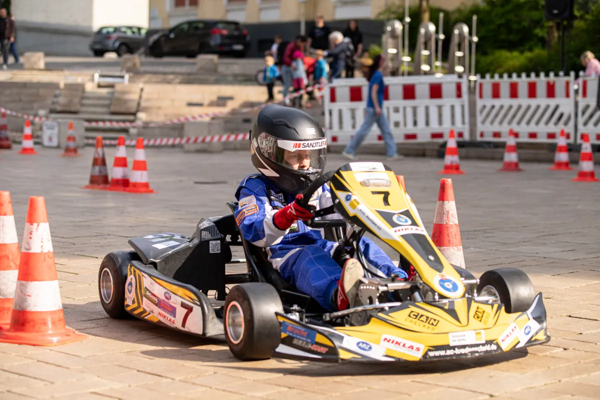 Ein Kartfahrer mit Helm und Rennanzug steuert ein gelb-blaues Kart auf einer abgesperrten Strecke durch die Innenstadt. Im Hintergrund sind Zuschauer und Verkehrshütchen zu sehen.