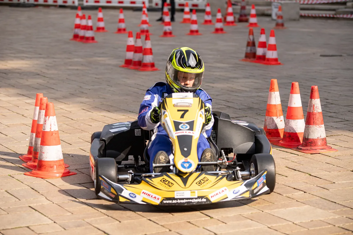 Ein Kartfahrer mit der Startnummer 7 steuert sein Fahrzeug auf einer abgesperrten Strecke zwischen orangefarbenen Pylonen durch die Innenstadt.