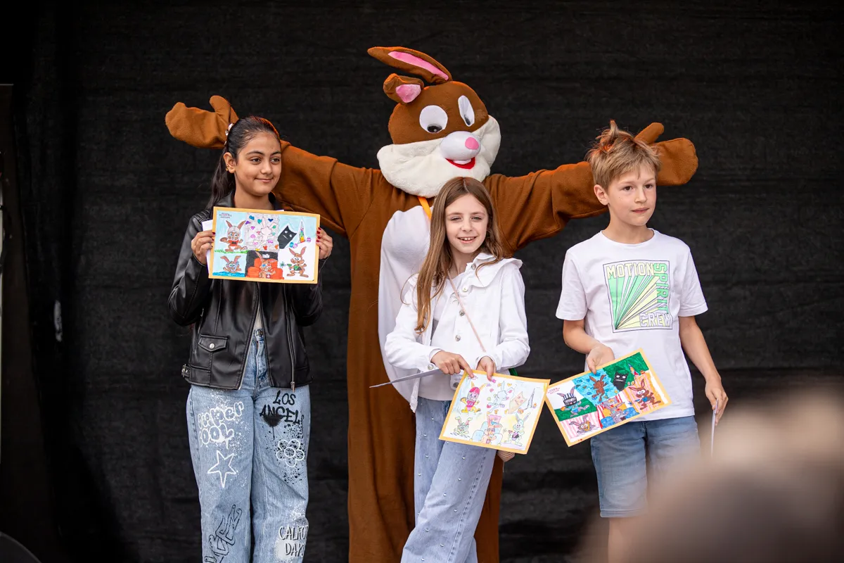 Drei Kinder halten bunte Zeichnungen vor sich, während ein großes Hasenkostüm hinter ihnen steht. Im Hintergrund ist ein dunkler Vorhang zu sehen.