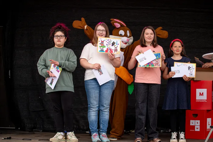 Vier Kinder halten auf einer Bühne Zeichnungen in den Händen, während ein großer, orangefarbener Hase neben ihnen steht. Im Hintergrund ist ein rotes Werbeplakat mit dem Buchstaben M zu sehen.