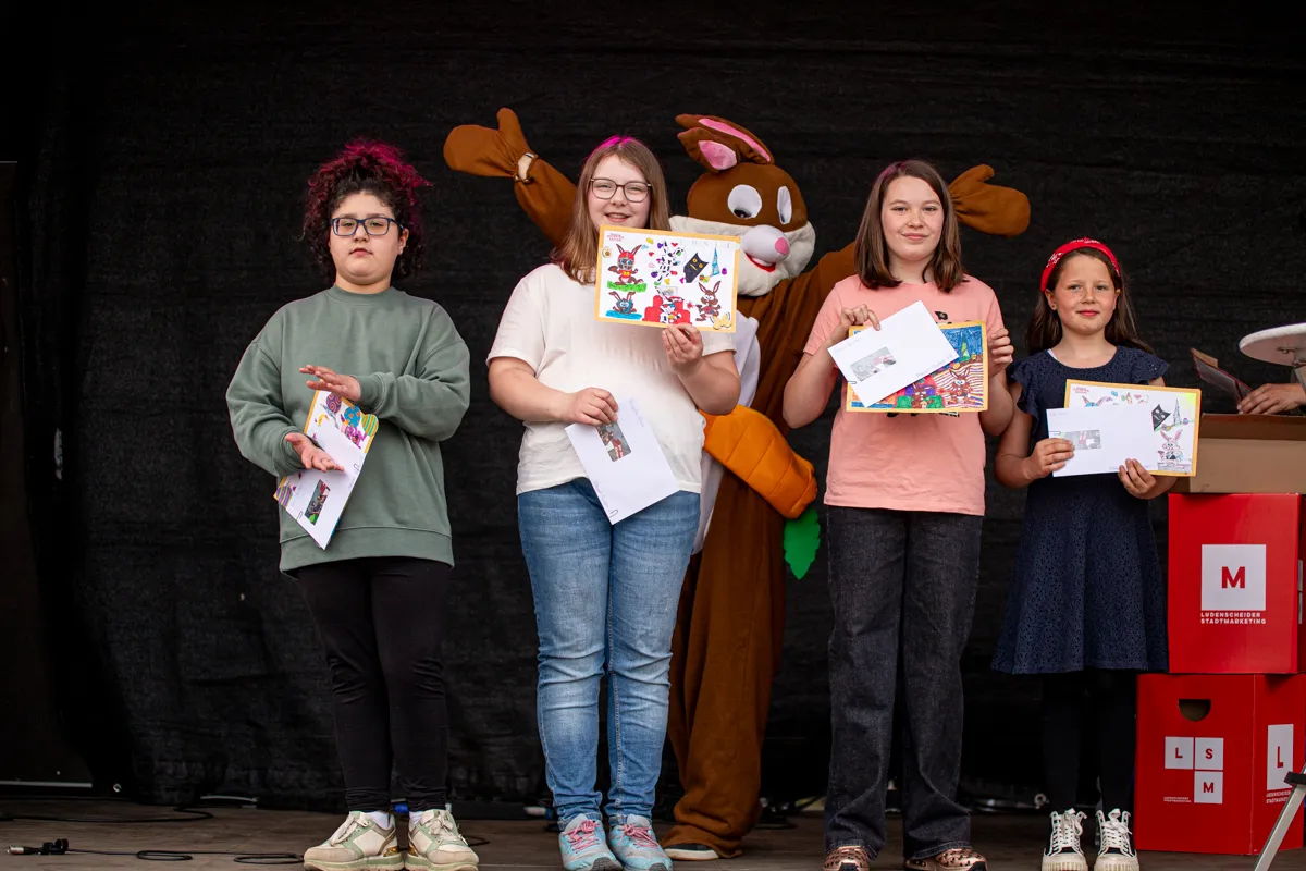 Vier Kinder halten auf einer Bühne Zeichnungen in den Händen, während ein großer, orangefarbener Hase neben ihnen steht. Im Hintergrund ist ein rotes Werbeplakat mit dem Buchstaben M zu sehen.