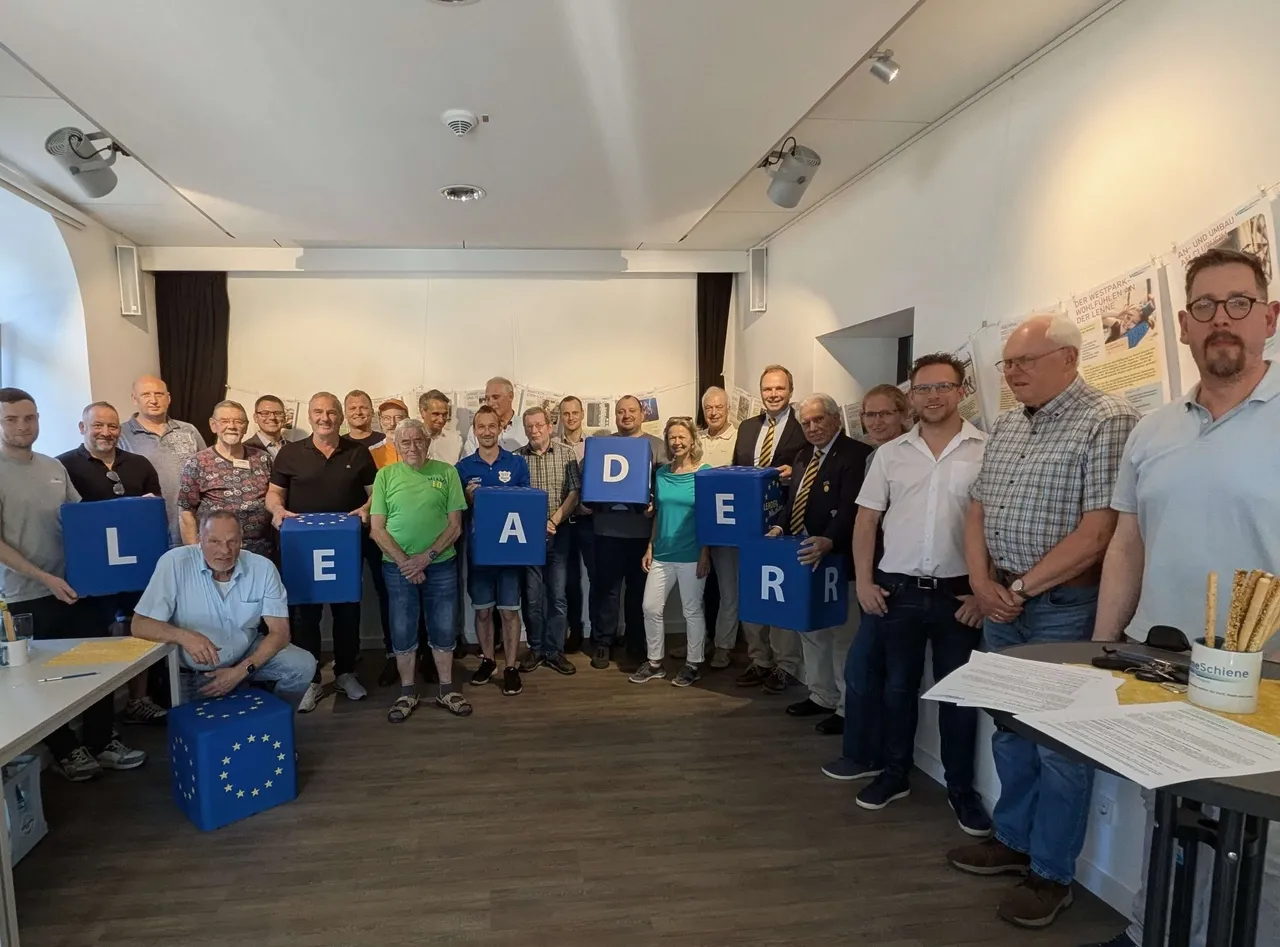 Eine Gruppe von etwa 20 Personen steht in einem Raum und hält große, farbige Buchstaben, die zusammen das Wort „LEADER“ ergeben. Im Hintergrund ist ein Plakat mit Text und Bildern zu sehen.