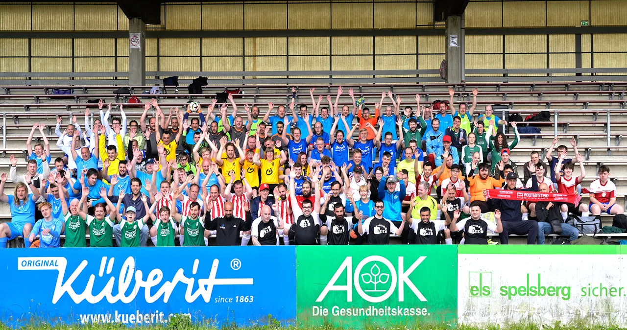 Eine große Gruppe von Menschen, hauptsächlich junge Männer, sitzen auf den Tribünen eines Stadions und heben die Arme in die Luft. Viele tragen Trikots in verschiedenen Farben, darunter Blau, Grün, Rot und Schwarz. Im Vordergrund sind Logos von Sponsoren wie Küberit, AOK und els Spelsberg zu sehen.