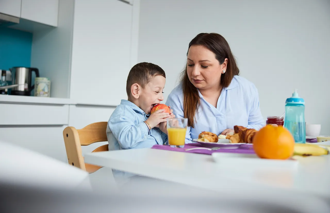 Eine Frau sitzt am Küchentisch und beobachtet, wie ihr kleiner Sohn in einen Apfel beißt. Vor ihnen stehen ein Glas Orangensaft, ein Croissant und Obst.