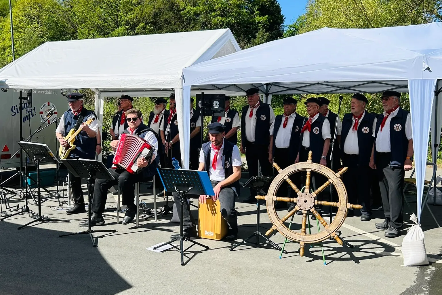 Musikalischer Auftritt eines Shanty-Chors unter einem weißen Zelt mit einem Steuerrad im Vordergrund.