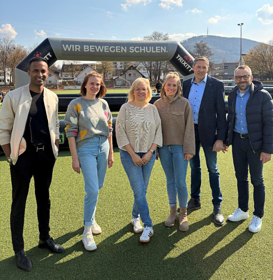 Sechs Personen stehen lächelnd auf einem grünen Sportplatz vor einem Banner mit der Aufschrift „Wir bewegen Schulen.“ im Hintergrund eine Hügellandschaft.