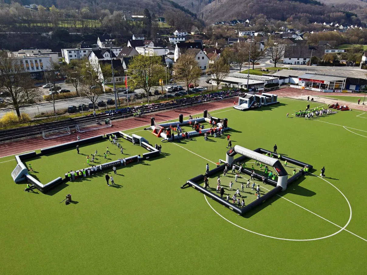 Luftaufnahme zeigt Kindergruppen, die auf einem Fußballfeld mit aufblasbaren Toren und Hindernissen spielen. Im Hintergrund sind Gebäude und ein Parkplatz zu sehen.
