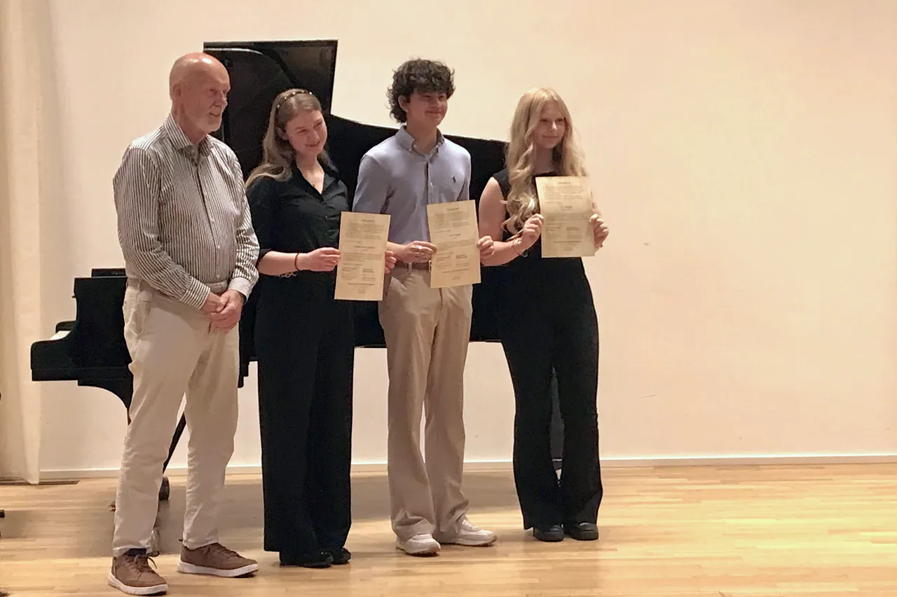 Nikolaus Goldbach steht mit den Musikpreisträgern Matilda Brandner, Josiah Santos und Lina Wiegold auf einer Bühne und hält Urkunden in der Hand. Im Hintergrund steht ein schwarzer Flügel.