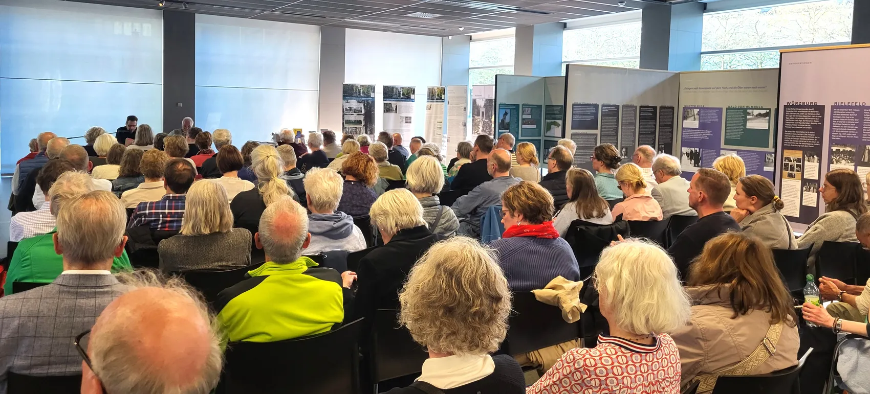 Zuhörer sitzen auf schwarzen Stühlen in einem hellen Raum und lauschen einem Mann, der am rechten Bildrand steht und spricht. Im Hintergrund hängen Poster mit historischen Bildern und Texten.