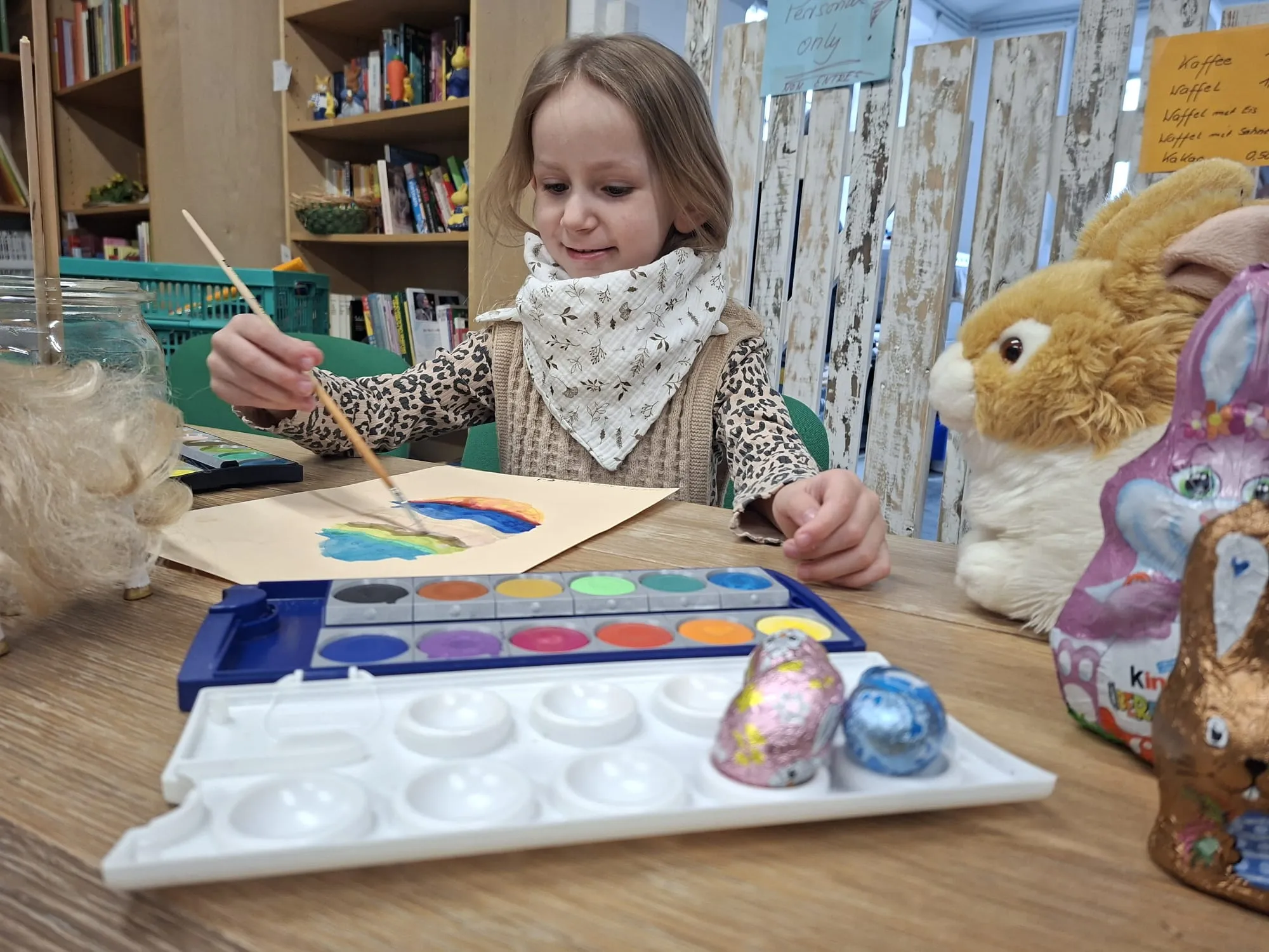 Ein Mädchen malt mit Wasserfarben an einem Tisch, umgeben von Osterhasen aus Plüsch und Schokolade.