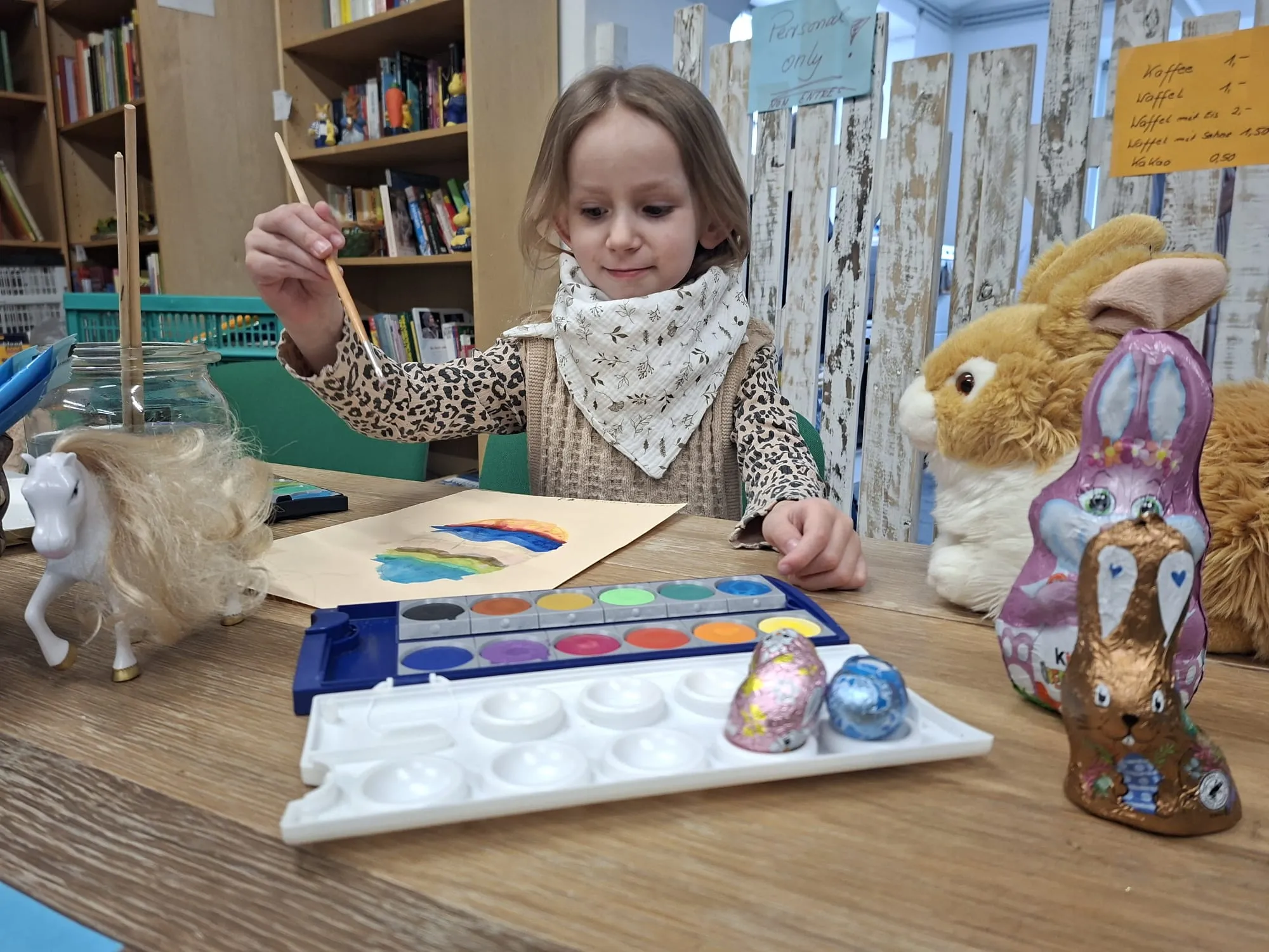 Ein Mädchen malt mit Wasserfarben an einem Tisch, vor dem mehrere Ostereier und Stoffhasen stehen.