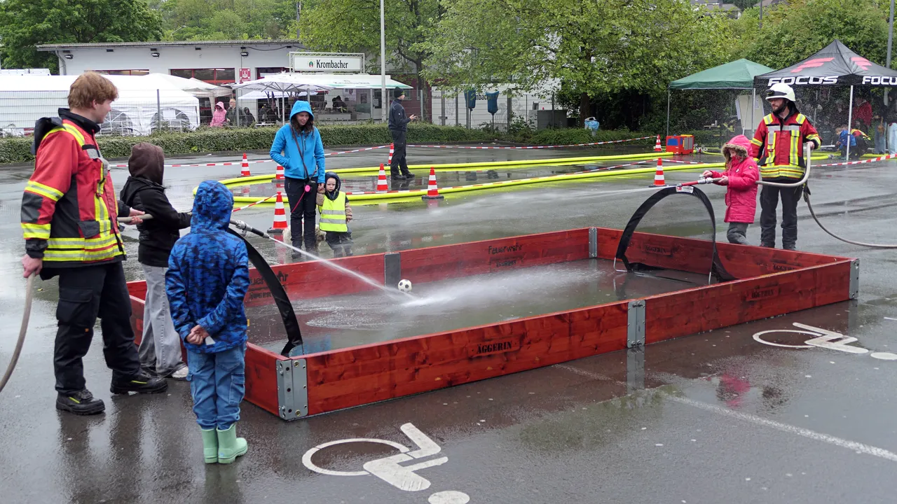 Mehrere Feuerwehrleute und Kinder stehen um eine mit Wasser gefüllte, rote Wanne. Ein Kind hält eine Wasserpistole und sprüht Wasser in die Wanne, während andere zusehen.