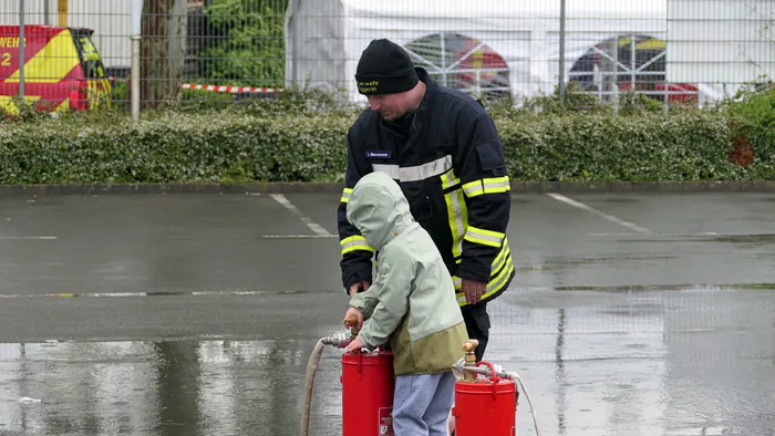 Ein Feuerwehrmann erklärt einem Kind die Bedienung eines roten Feuerlöschers auf einem nassen Parkplatz.