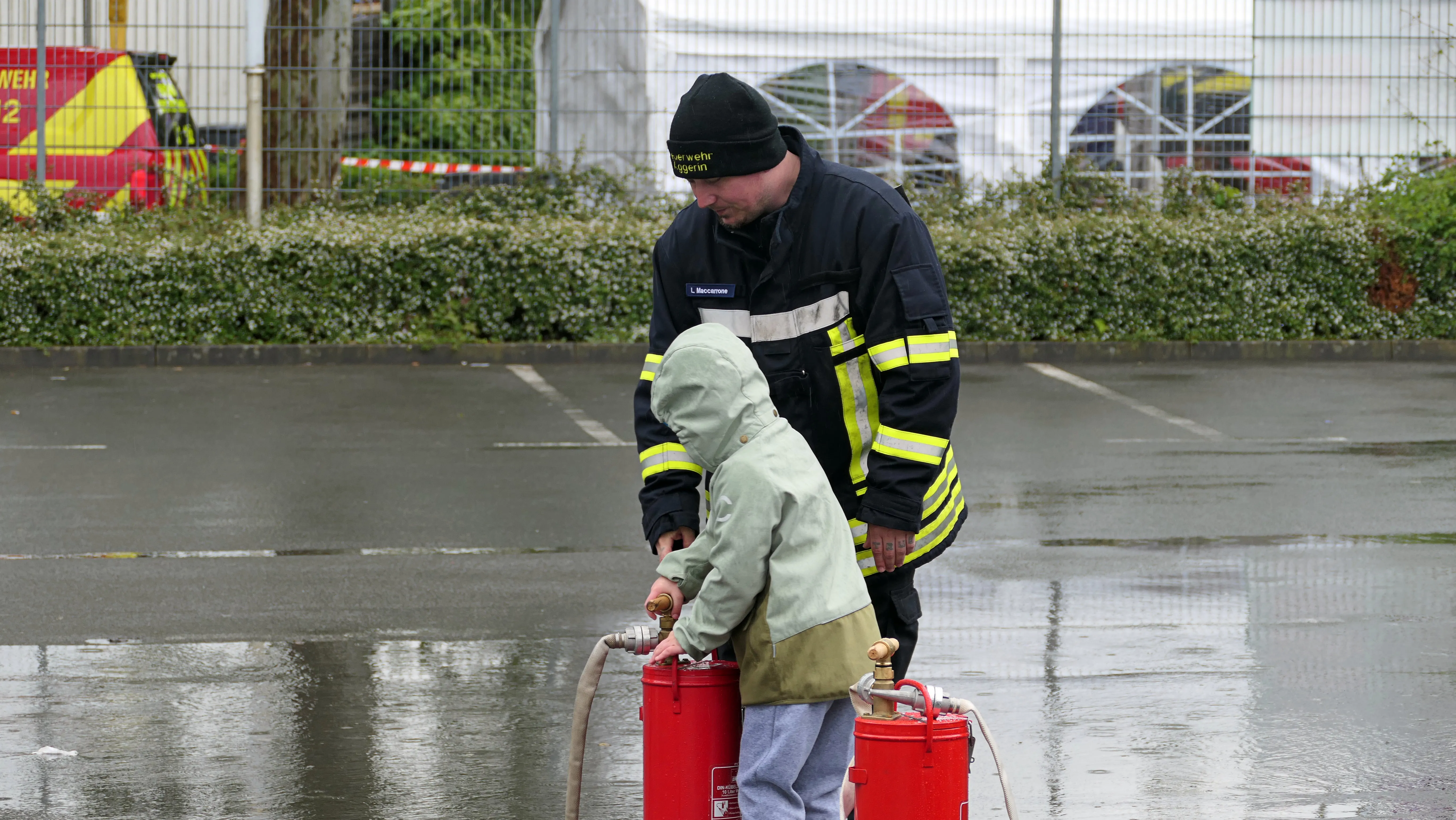 Ein Feuerwehrmann erklärt einem Kind die Bedienung eines roten Feuerlöschers auf einem nassen Parkplatz.