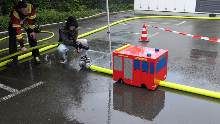 Zwei Personen beobachten ein ferngesteuertes Modell eines Feuerwehrautos auf einem nassen Parkplatz, umgeben von Feuerwehrschläuchen.