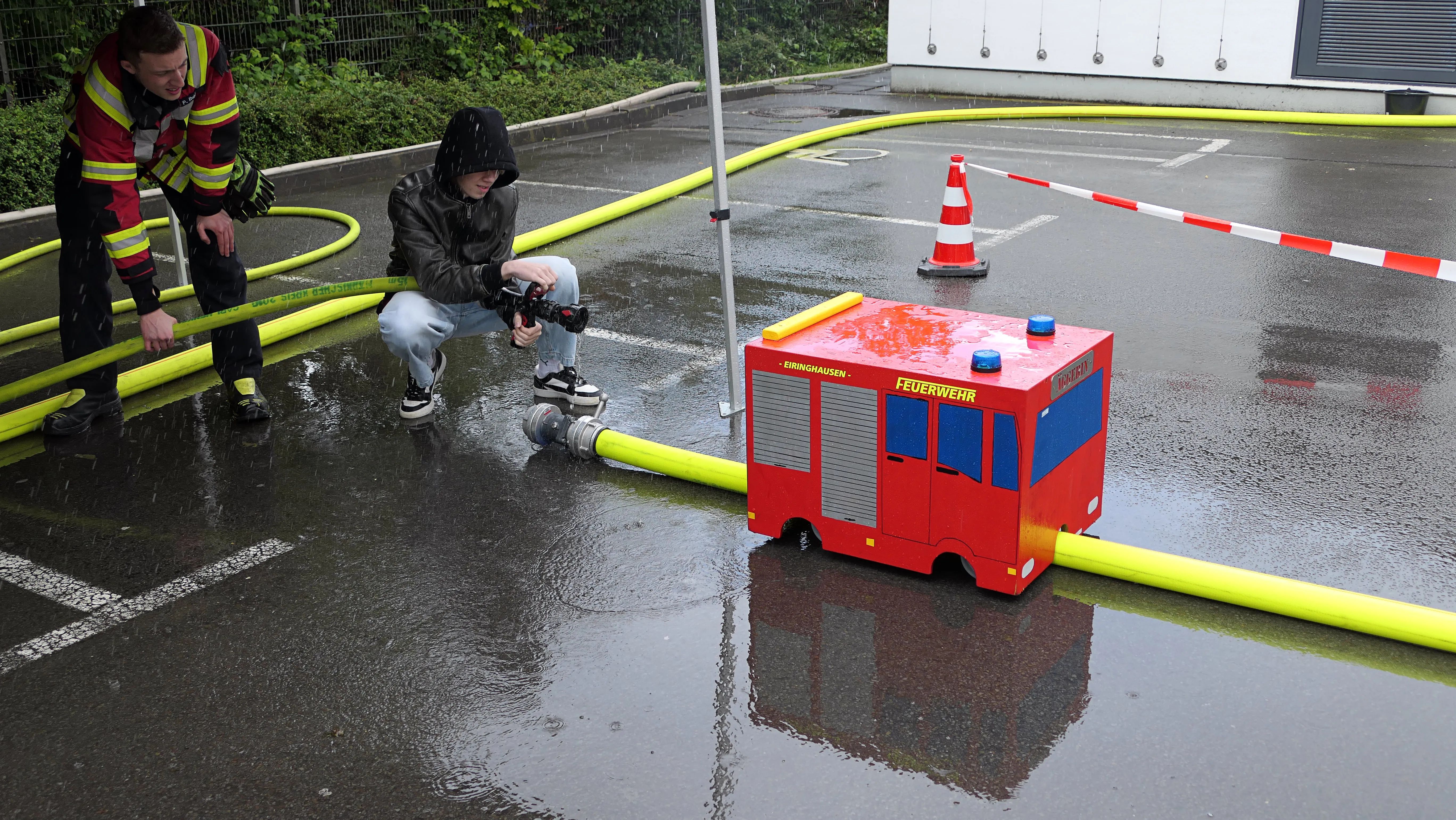 Zwei Personen beobachten ein ferngesteuertes Modell eines Feuerwehrautos auf einem nassen Parkplatz, umgeben von Feuerwehrschläuchen.
