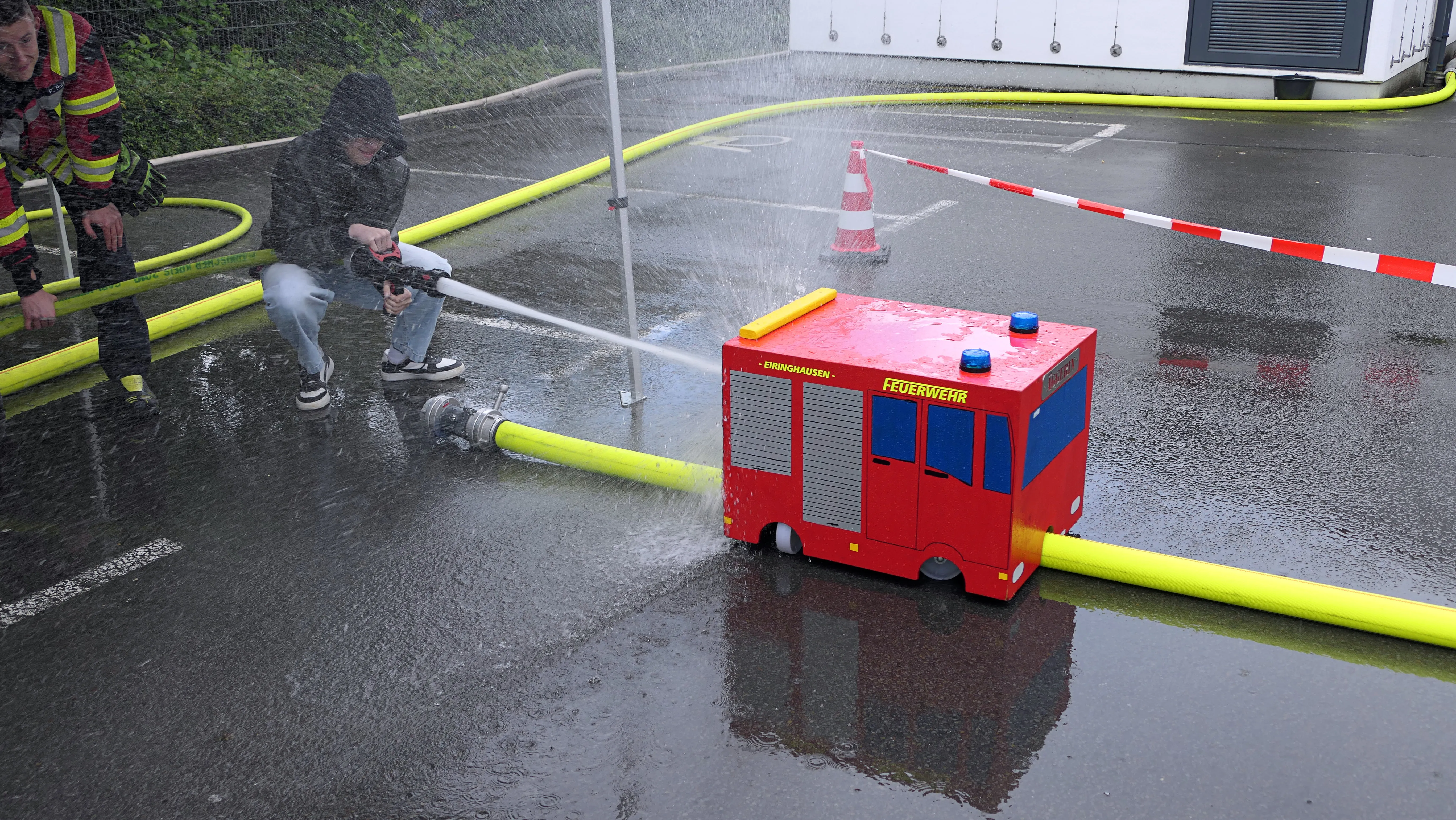 Ein rotes Spielzeug-Feuerwehrfahrzeug wird von einem Wasserstrahl aus einem Schlauch getroffen, der von einer Person gehalten wird. Im Hintergrund steht ein Absperrgitter und ein Gebäude.