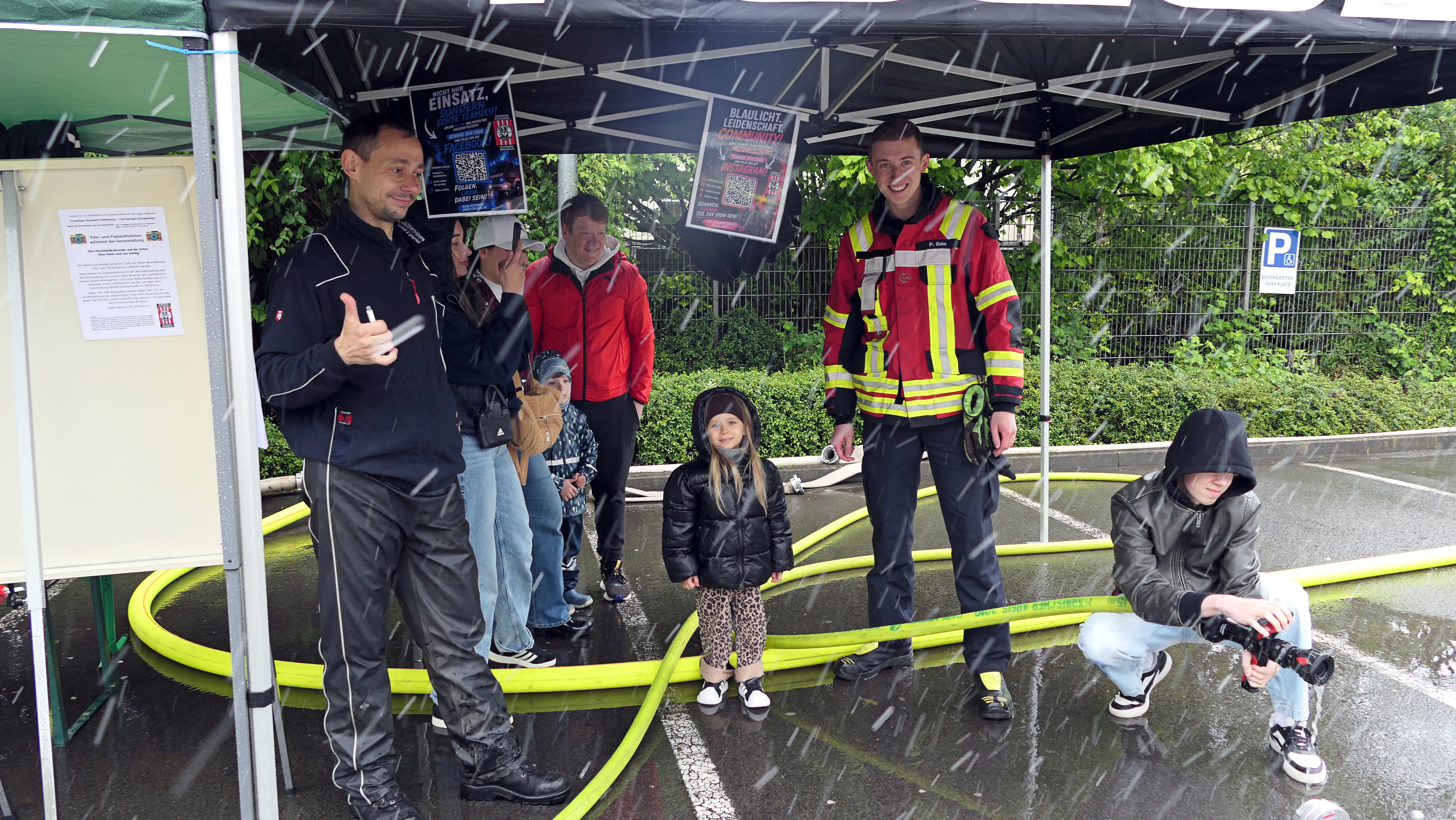 Feuerwehrleute und Besucher stehen lachend unter einem Zelt im Regen, während ein Kind mit einem Wasserschlauch spielt. Im Hintergrund sind Plakate und Bäume zu sehen.
