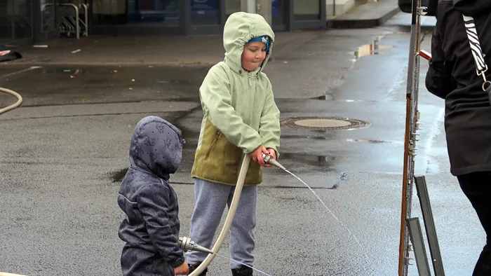Zwei Kinder üben Bogenschießen unter Aufsicht eines Erwachsenen im Freien. Der ältere Junge zielt mit dem Bogen, während der jüngere zusieht.