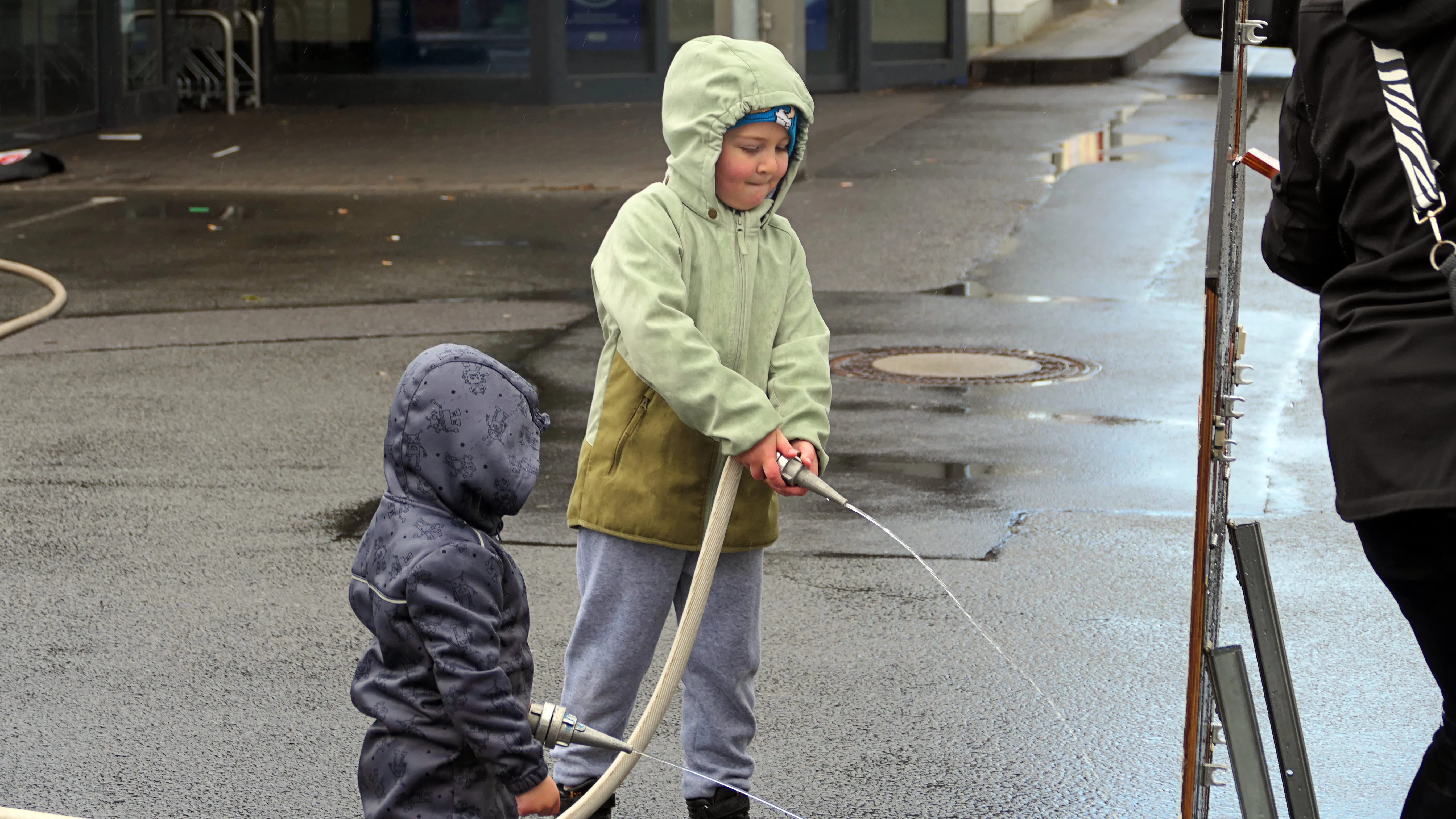 Zwei Kinder üben Bogenschießen unter Aufsicht eines Erwachsenen im Freien. Der ältere Junge zielt mit dem Bogen, während der jüngere zusieht.