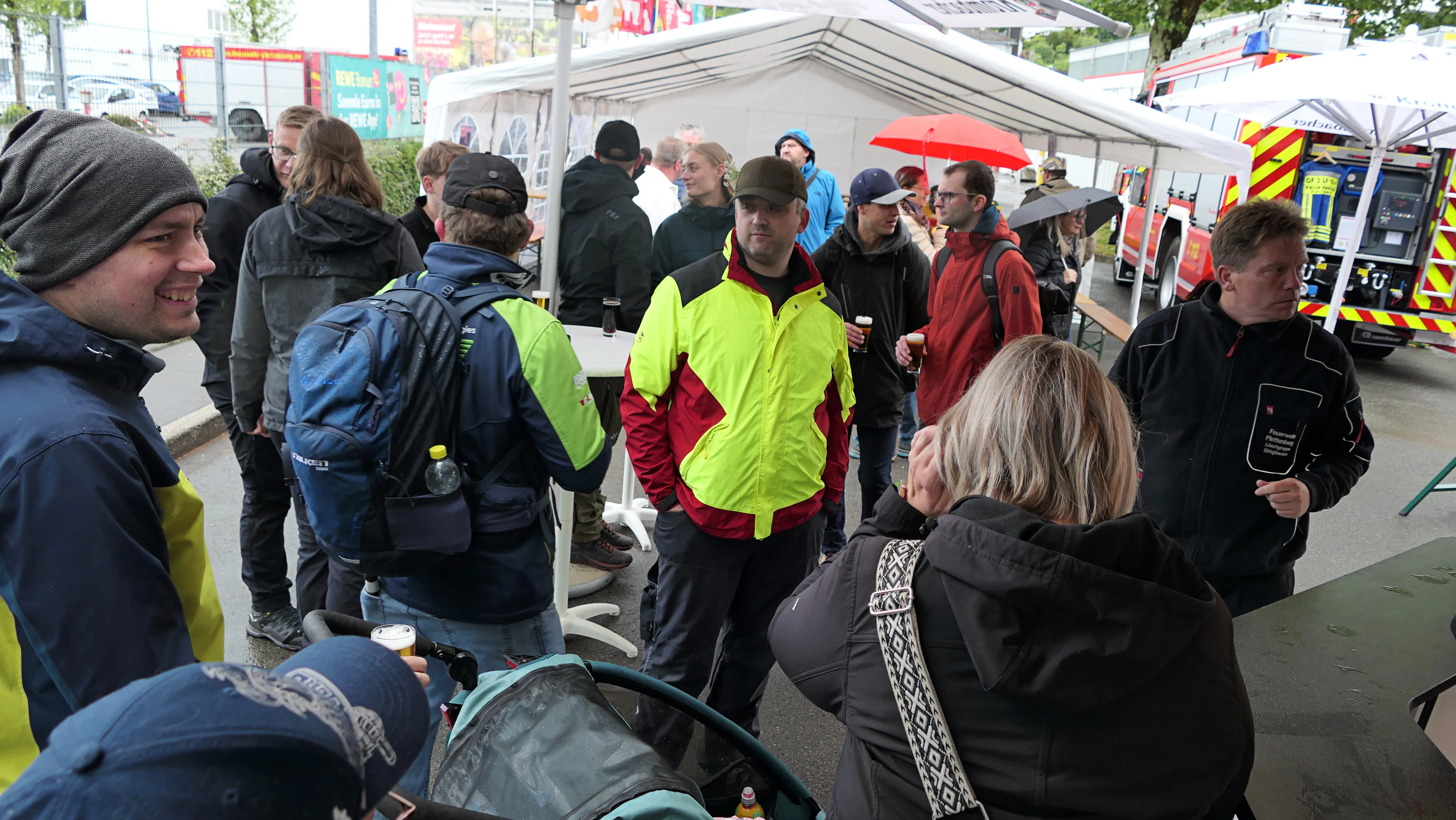 Menschengruppen stehen und unterhalten sich auf einem Festgelände mit Zelten und einem Feuerwehrauto im Hintergrund.