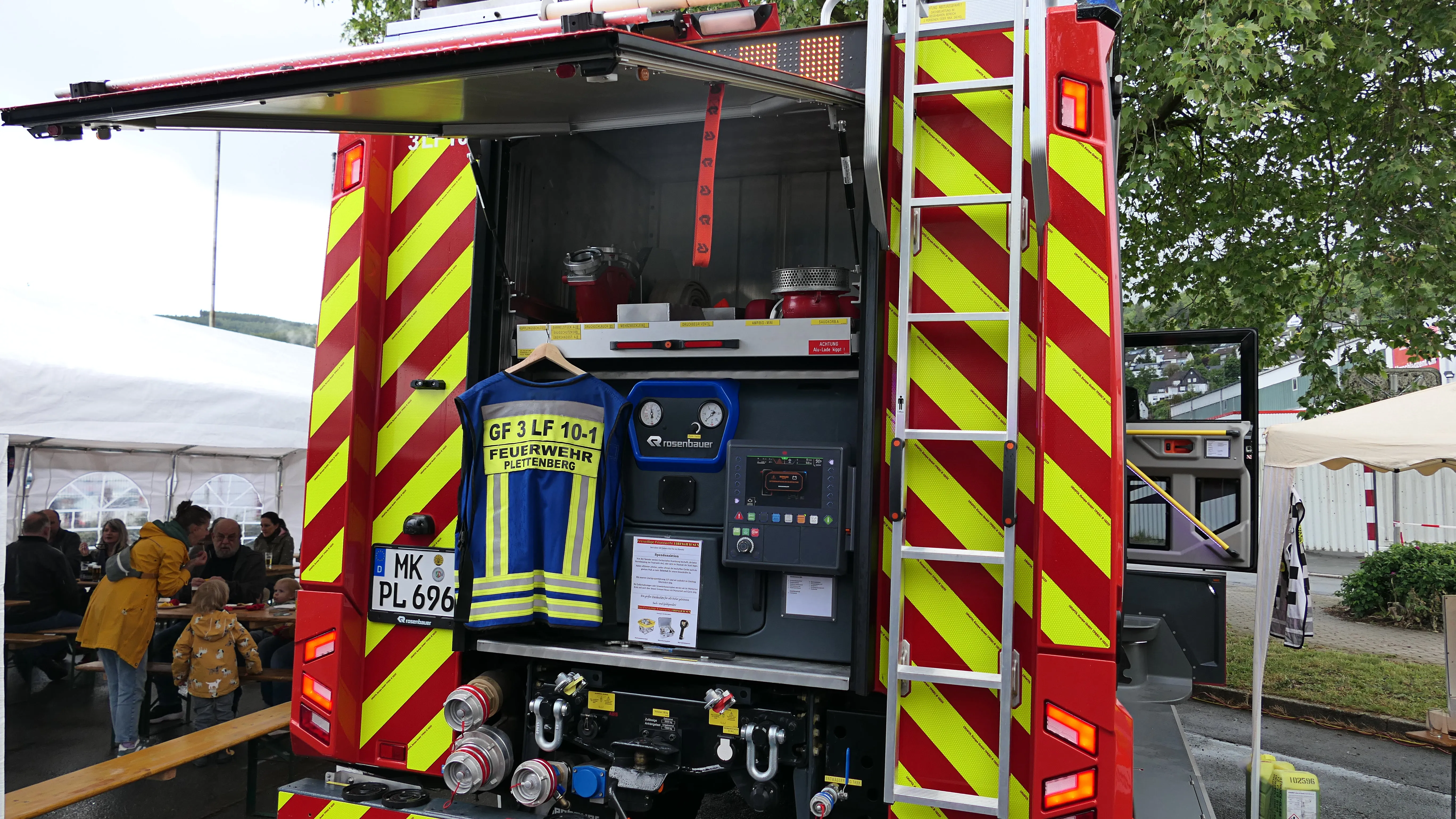Heckansicht eines roten Feuerwehrautos mit gelben Streifen, an dem eine Feuerwehruniform hängt. Im Hintergrund sind Menschen und ein Zelt zu sehen.