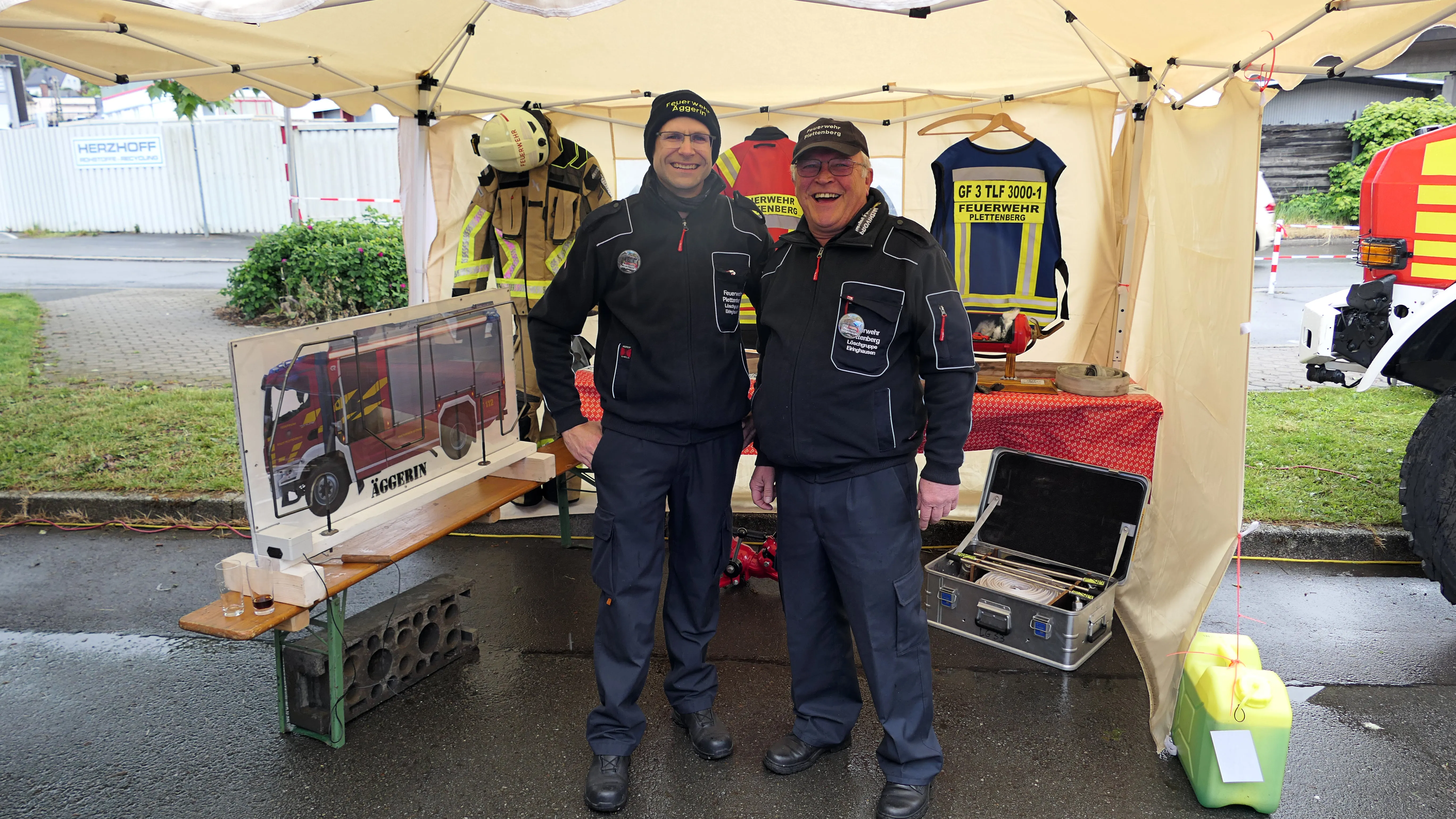 Zwei Männer in Uniformen der Freiwilligen Feuerwehr stehen vor einem Zelt mit Ausrüstung und einem Modell eines Feuerwehrautos. Im Hintergrund ist ein rotes Feuerwehrfahrzeug zu sehen.