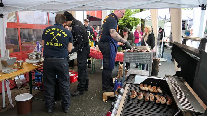 Feuerwehrleute grillen Würstchen an einem Stand mit dem Schriftzug „Feuerwehr Äggerin“ für Besucher eines Familientags.