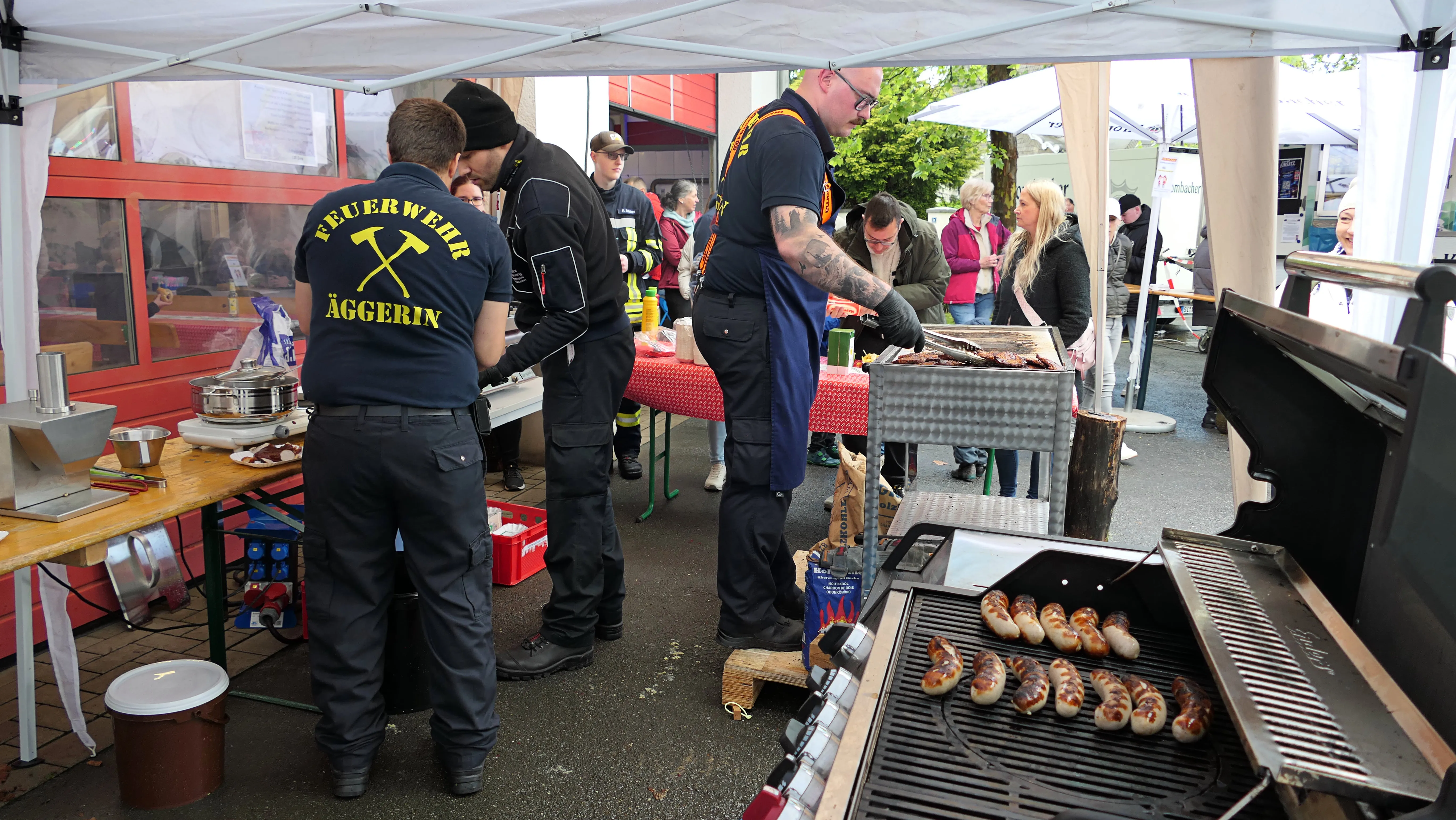 Feuerwehrleute grillen Würstchen an einem Stand mit dem Schriftzug „Feuerwehr Äggerin“ für Besucher eines Familientags.