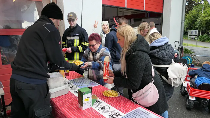 Menschen stehen an einem Tisch mit Pommes und Soßen, während im Hintergrund Feuerwehrleute stehen. Im Hintergrund ist ein rotes Feuerwehrauto zu sehen.