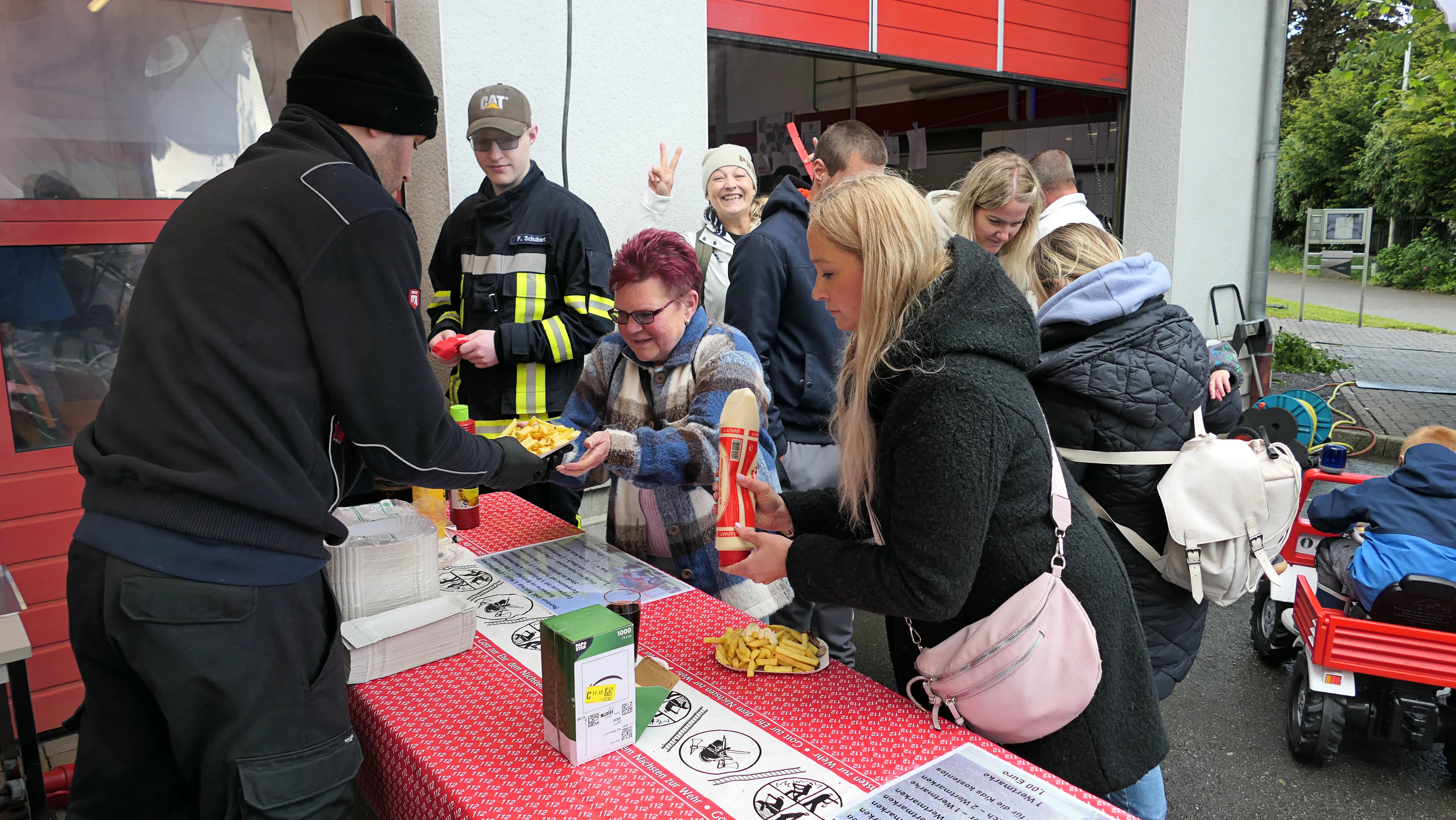 Menschen stehen an einem Tisch mit Pommes und Soßen, während im Hintergrund Feuerwehrleute stehen. Im Hintergrund ist ein rotes Feuerwehrauto zu sehen.