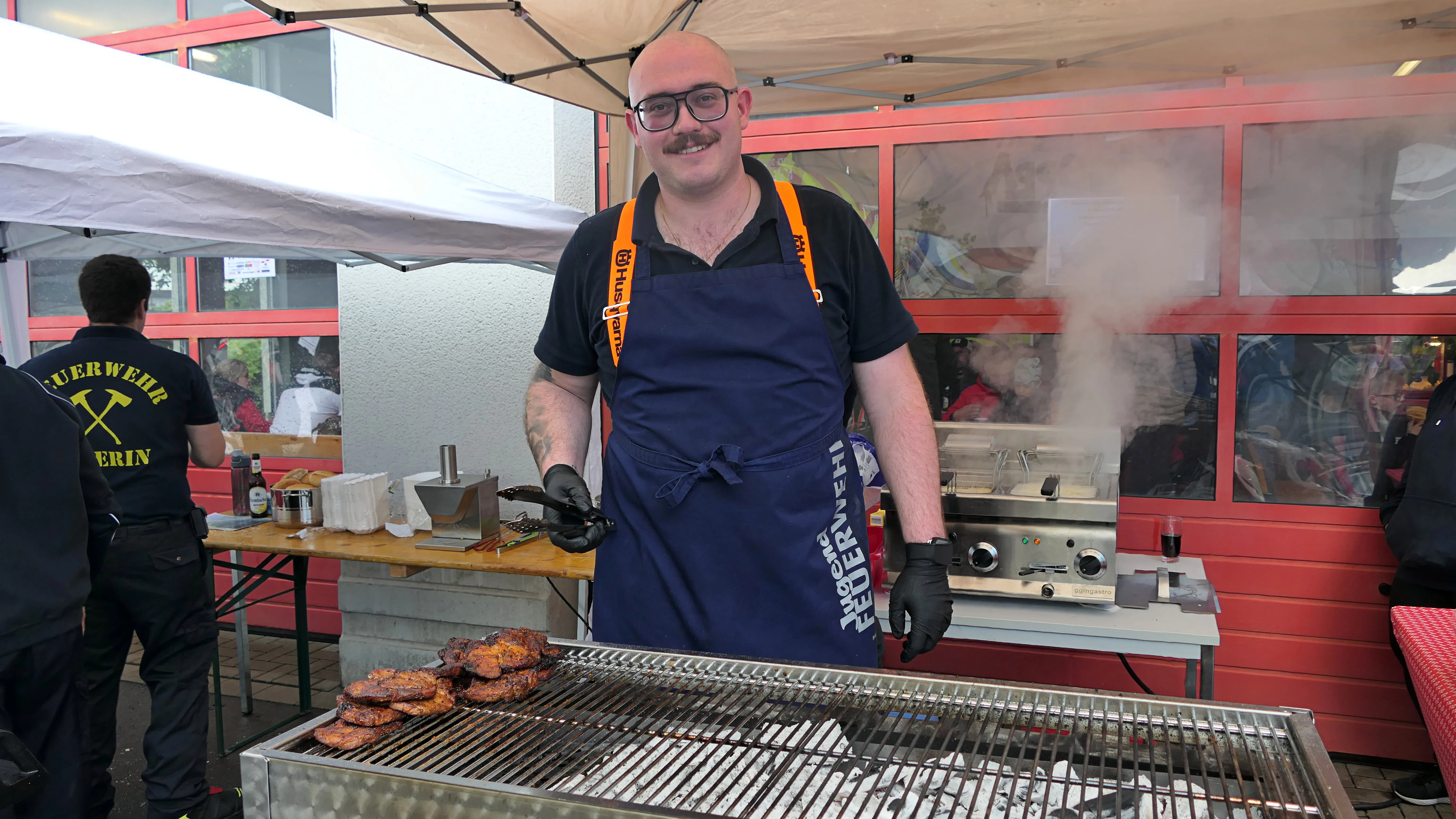 Ein Mann mit Glatze und Schnurrbart steht lächelnd vor einem Grill und brät Fleischstücke. Er trägt eine blaue Schürze mit dem Aufdruck „Freiwillige Feuerwehr“ und eine orangefarbene Tragehilfe. Im Hintergrund ist ein Zelt und ein Feuerwehrmann zu sehen.
