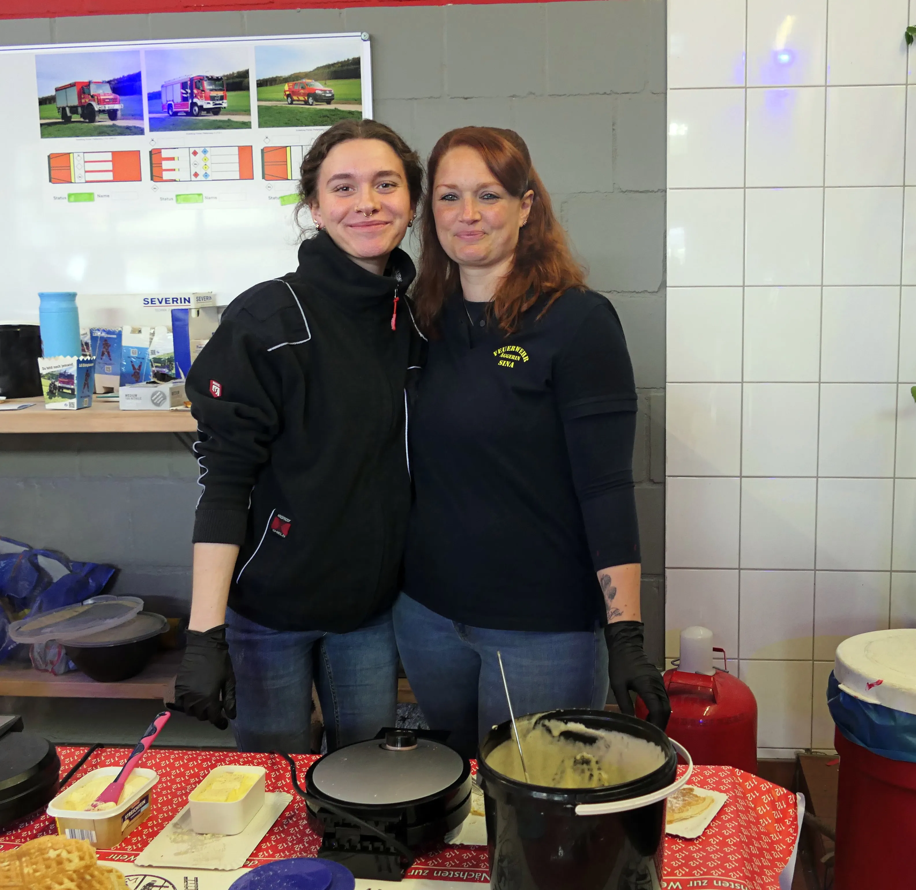 Zwei Frauen stehen hinter einem Tisch mit Speisen und bereiten etwas zu. Im Hintergrund sind ein Bild von Feuerwehrfahrzeugen und Geräte zu sehen.