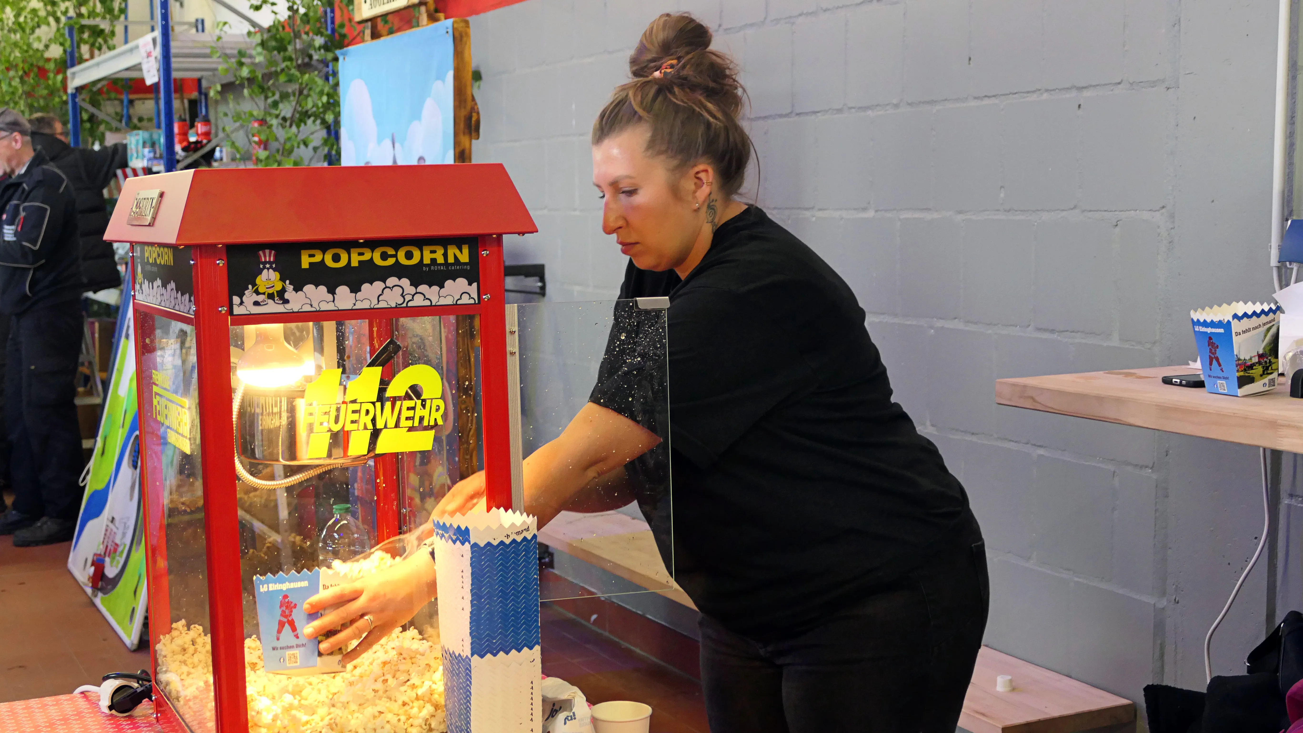 Eine Frau füllt Popcorn aus einer Maschine in einen Pappbecher. Im Hintergrund ist ein Stand mit roten und weißen Elementen zu sehen.