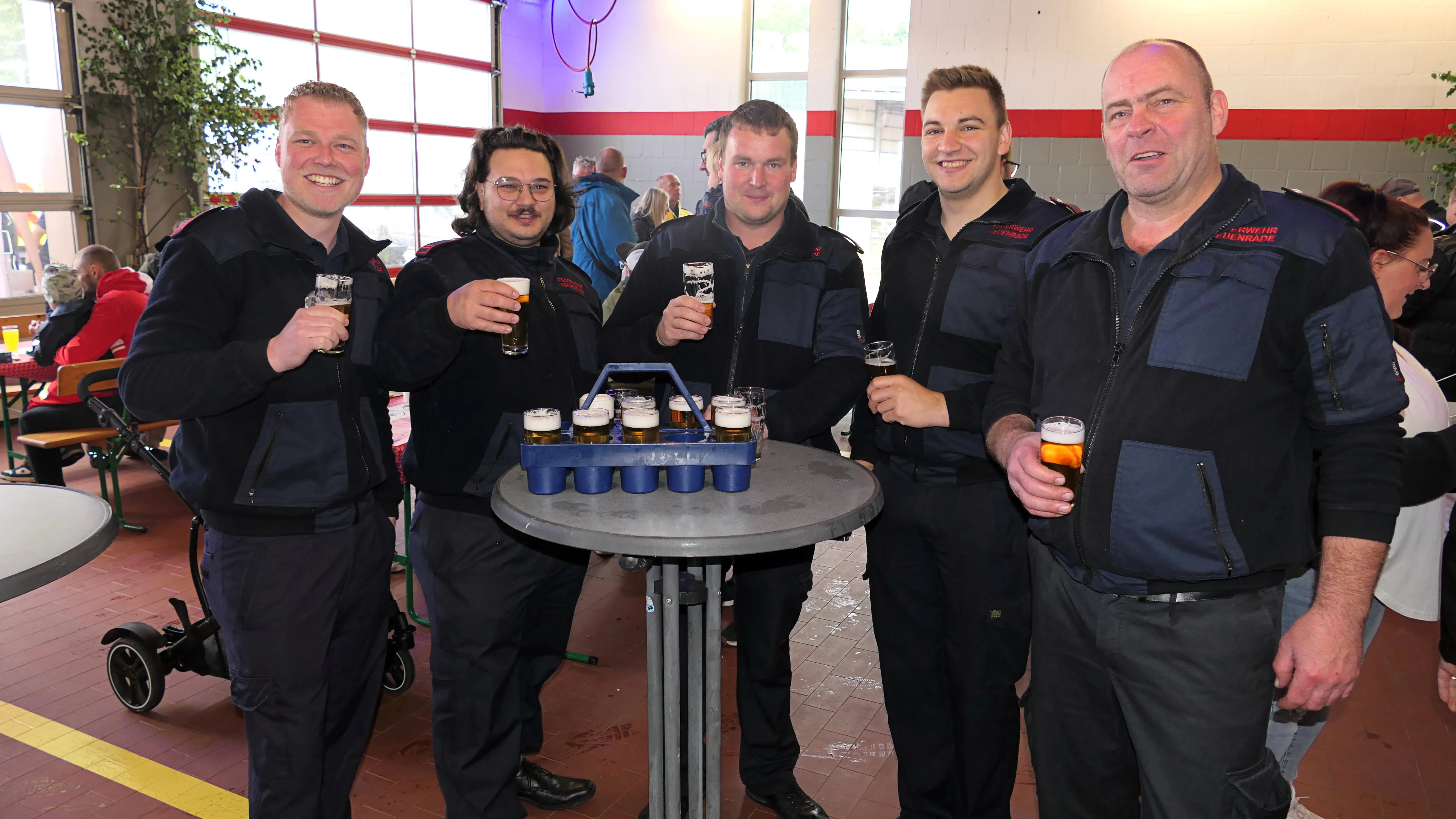Fünf Feuerwehrleute in Uniformen stehen um einen runden Tisch mit gefüllten Biergläsern und einer Getränkekiste. Im Hintergrund sind weitere Menschen und eine offene Tür zu sehen.