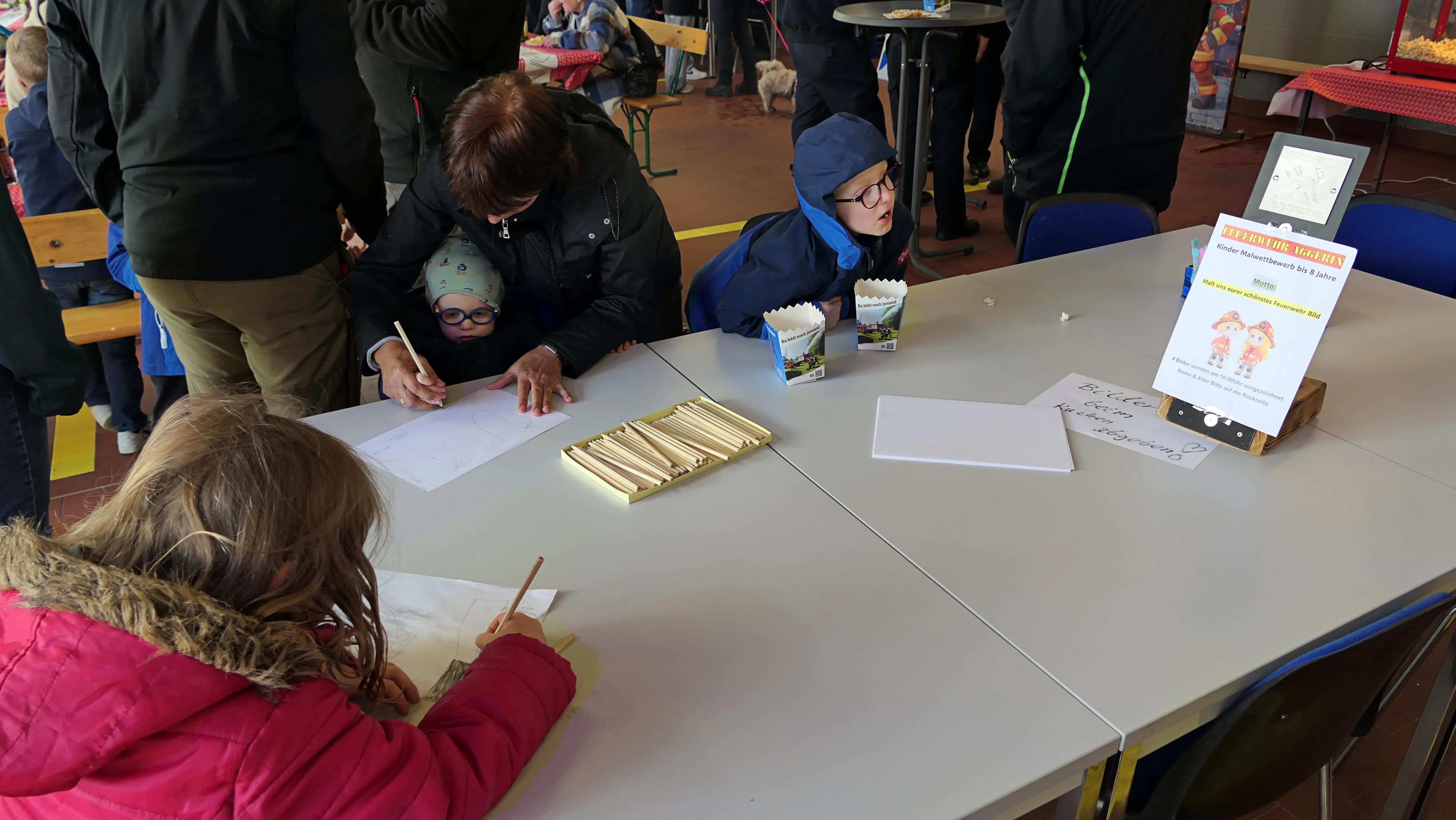 Kinder sitzen an einem Tisch und basteln oder malen auf einem Familientag der Löschgruppe Eiringhausen. Im Hintergrund ein Plakat mit dem Hinweis auf ein Angebot für Kinder zwischen drei und acht Jahren.