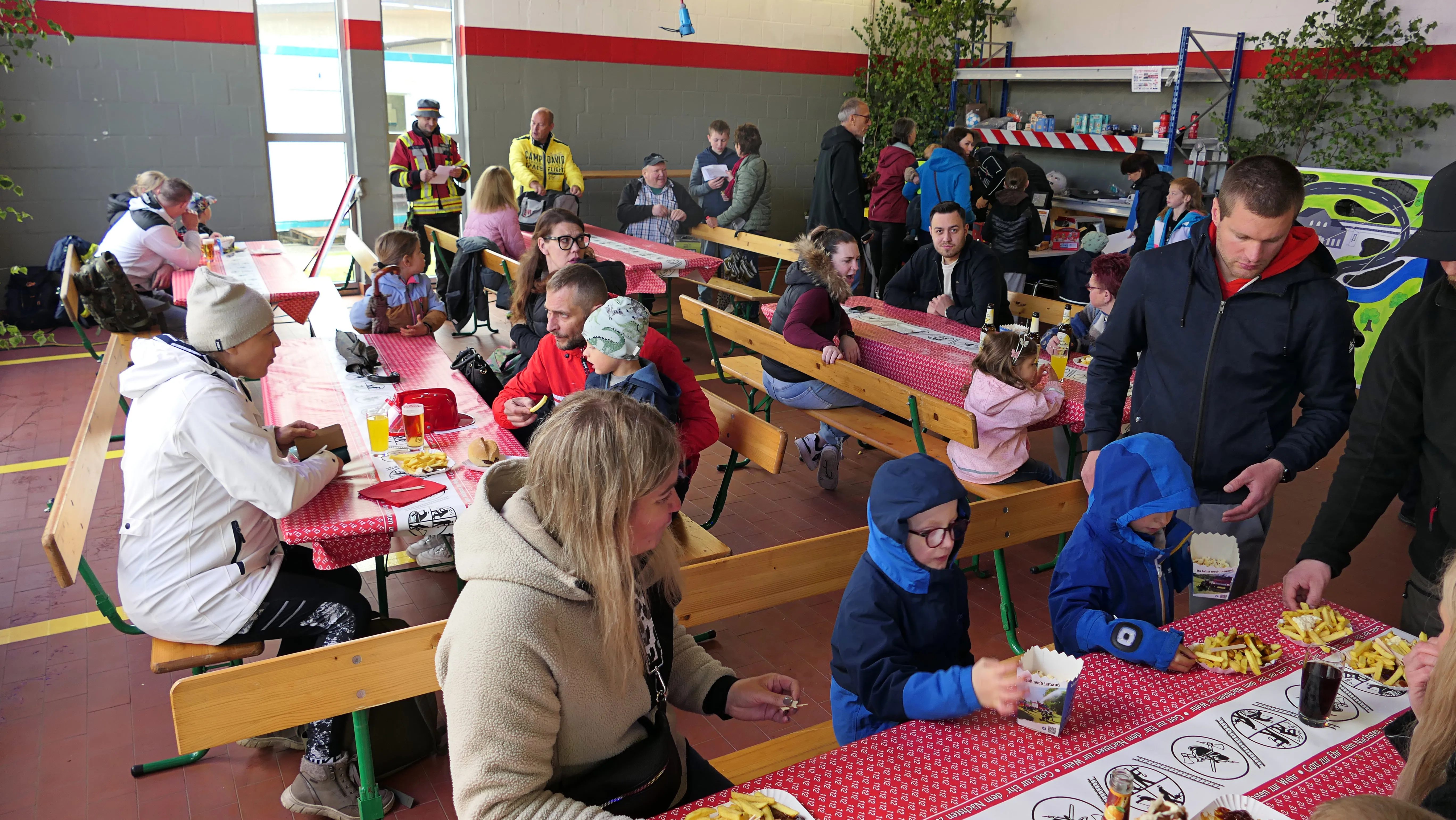 Menschen jeden Alters sitzen an Tischen in einer Turnhalle und essen Pommes und andere Speisen. Im Hintergrund sind weitere Personen und ein Verpflegungsstand zu sehen.