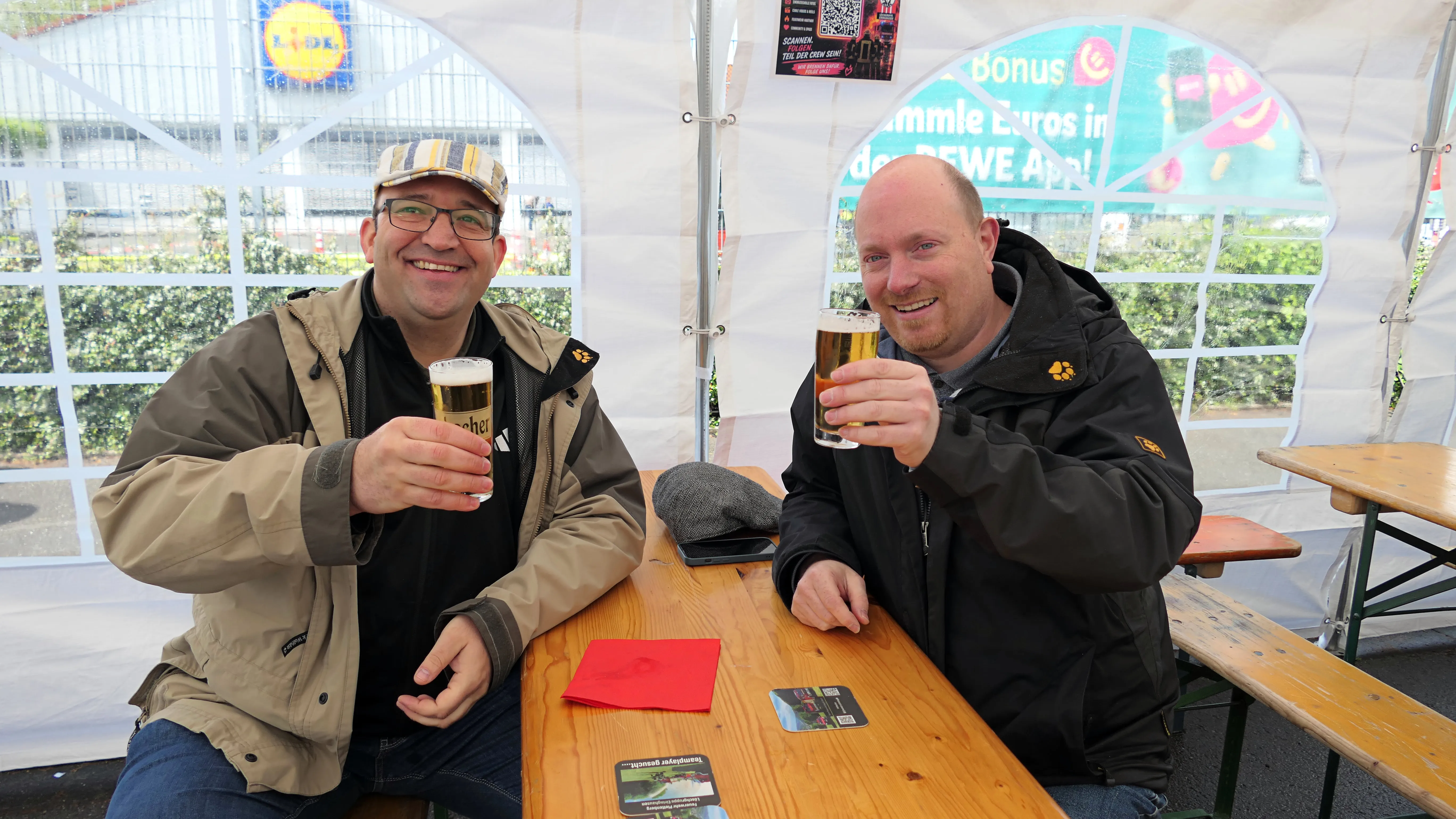 Zwei Männer sitzen an einem Holztisch unter einem Zelt und halten jeweils ein Bierglas hoch. Im Hintergrund ist ein Werbebanner zu sehen.