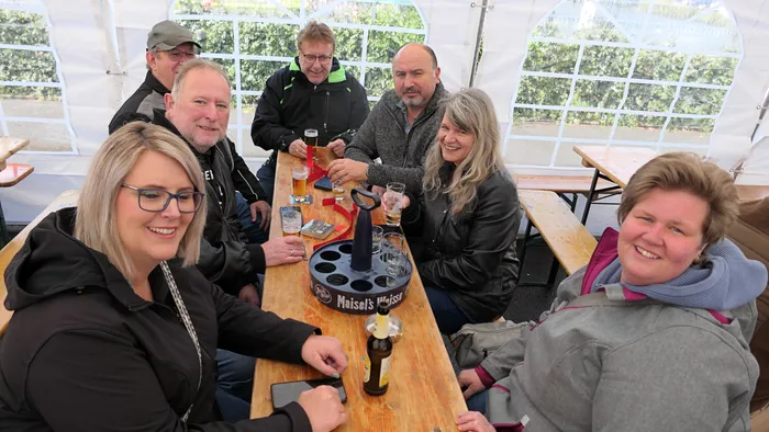 Eine Gruppe von sechs lächelnden Erwachsenen sitzt an einem Holztisch unter einem Zelt und genießt Getränke. In der Mitte steht ein Eiskübel mit Maisel's Weisse Bier.