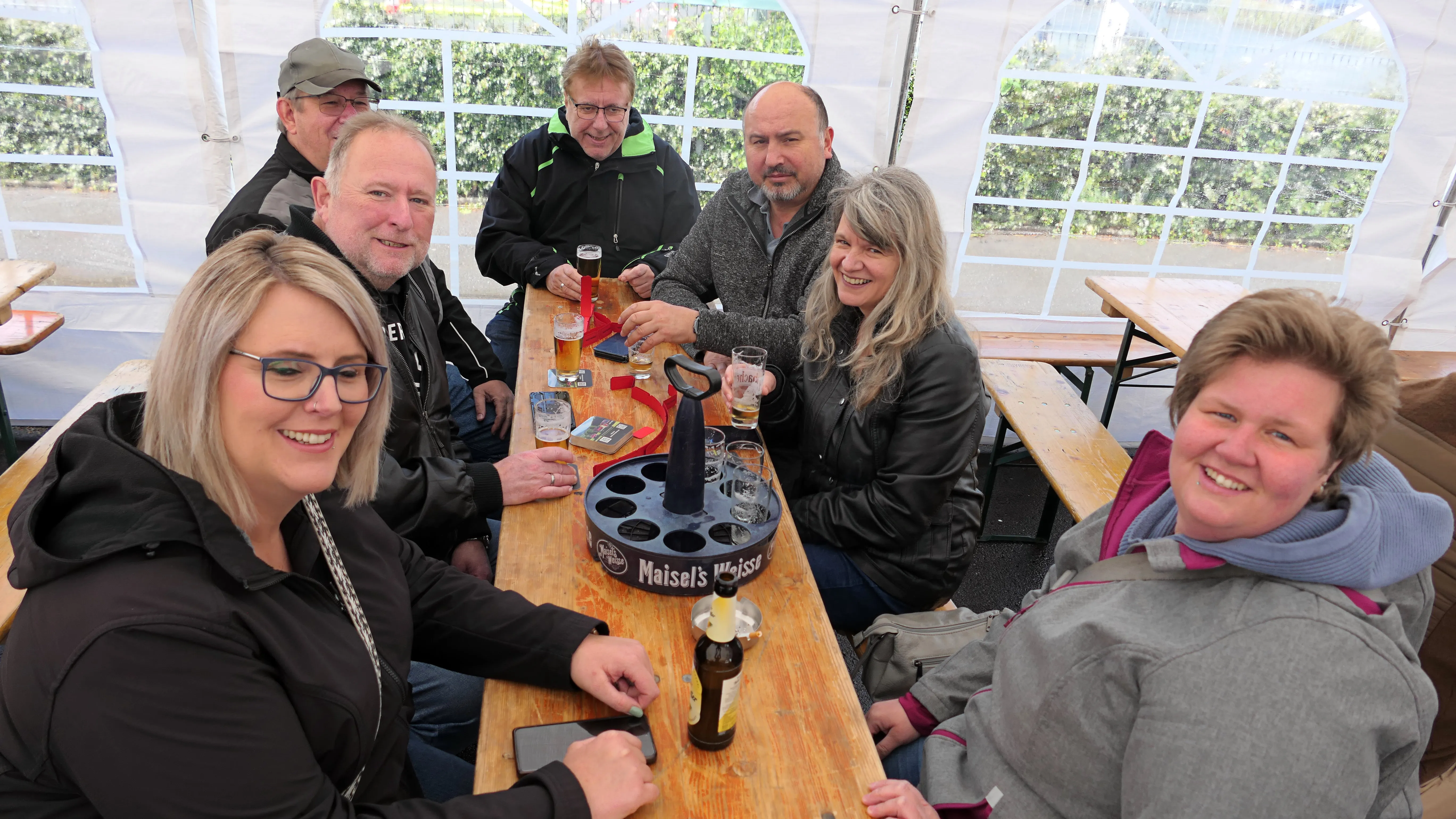 Eine Gruppe von sechs lächelnden Erwachsenen sitzt an einem Holztisch unter einem Zelt und genießt Getränke. In der Mitte steht ein Eiskübel mit Maisel's Weisse Bier.