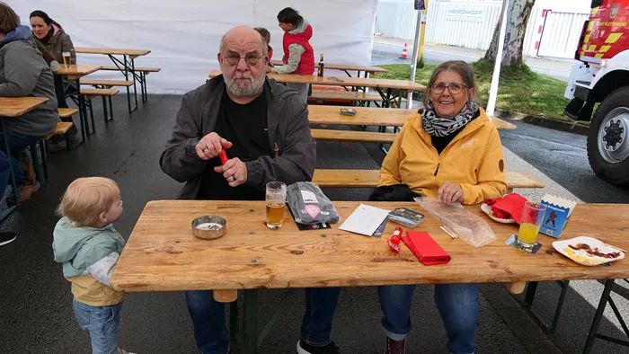 Ein älteres Paar sitzt an einem Biergartentisch mit einem kleinen Kind. Vor ihnen stehen Getränke und Snacks, im Hintergrund weitere Besucher eines Festes.