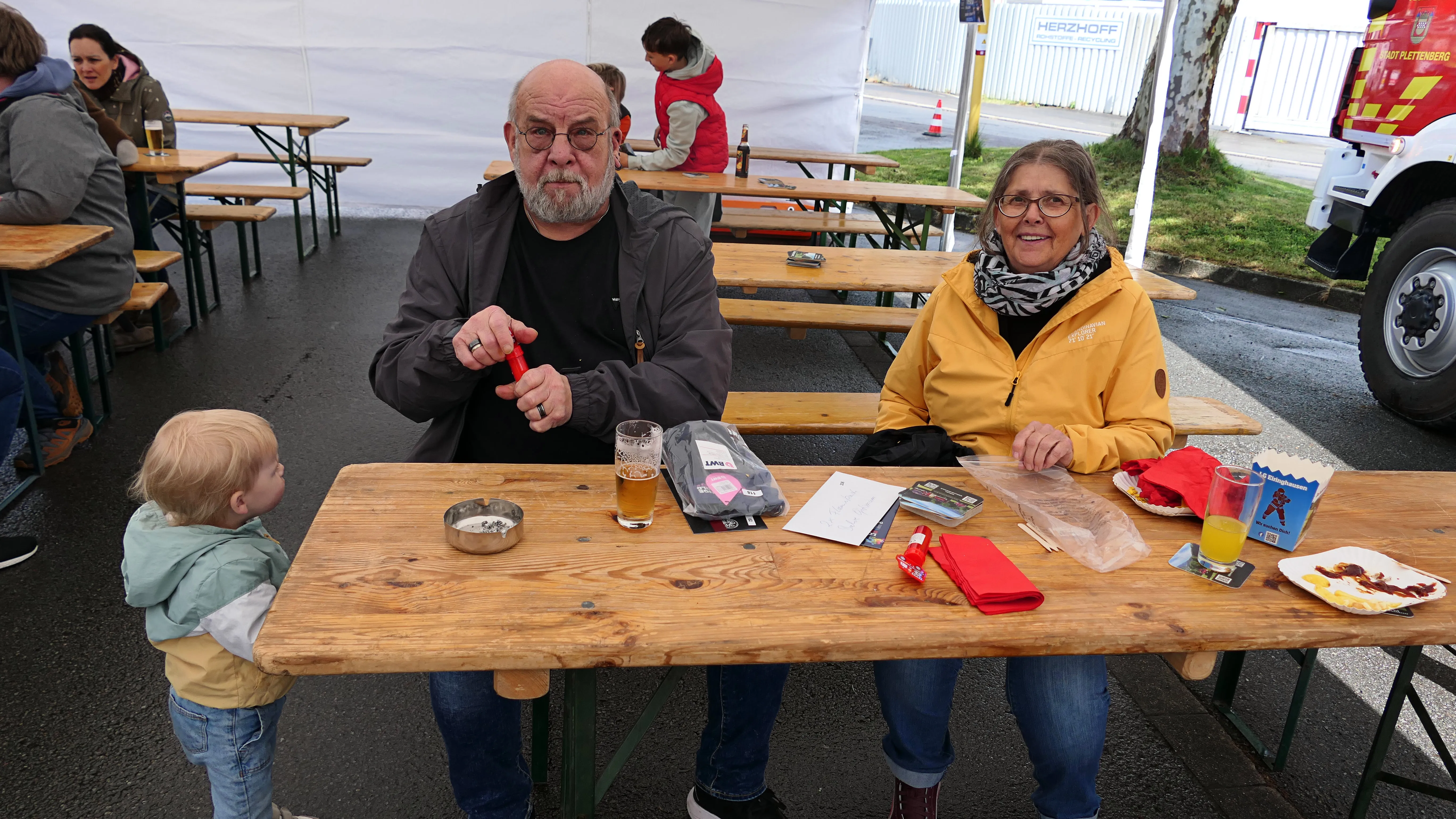 Ein älteres Paar sitzt an einem Biergartentisch mit einem kleinen Kind. Vor ihnen stehen Getränke und Snacks, im Hintergrund weitere Besucher eines Festes.