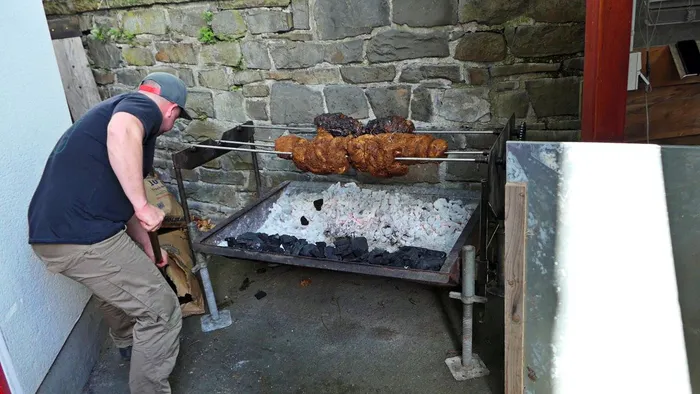 Ein Mann trägt Holzkohle zu einem Grill, auf dem mehrere Fleischstücke aufgespießt und gebraten werden. Im Hintergrund eine Steinmauer und ein Teil eines Gebäudes.