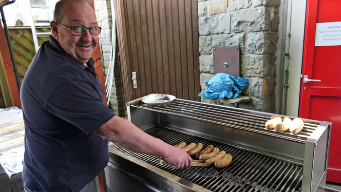 Ein Mann mit Brille und rotem Hemd steht lächelnd vor einem Grill und wendet Würstchen. Im Hintergrund ein roter Türrahmen und ein Schild an einer Steinmauer.