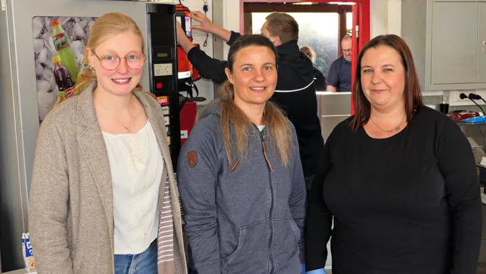 Drei Frauen stehen lächelnd vor einem Getränkeautomaten und einem roten Schrank in einem Raum mit einem Tresen im Hintergrund.