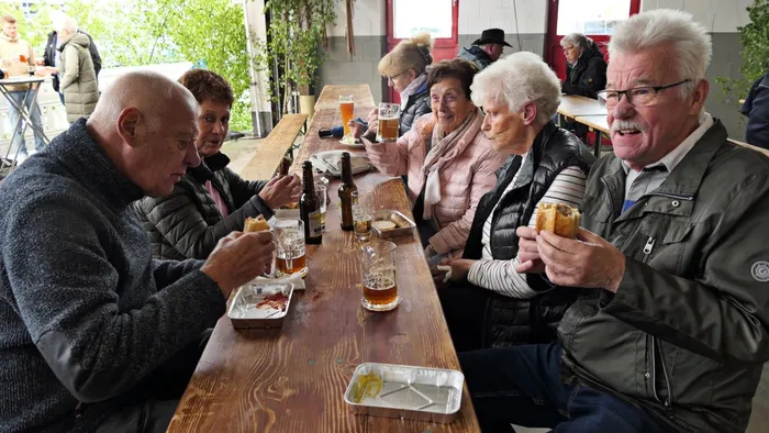 Mehrere ältere Menschen sitzen an einem langen Holztisch im Freien, essen und trinken zusammen. Im Hintergrund weitere Personen und ein Gebäude.