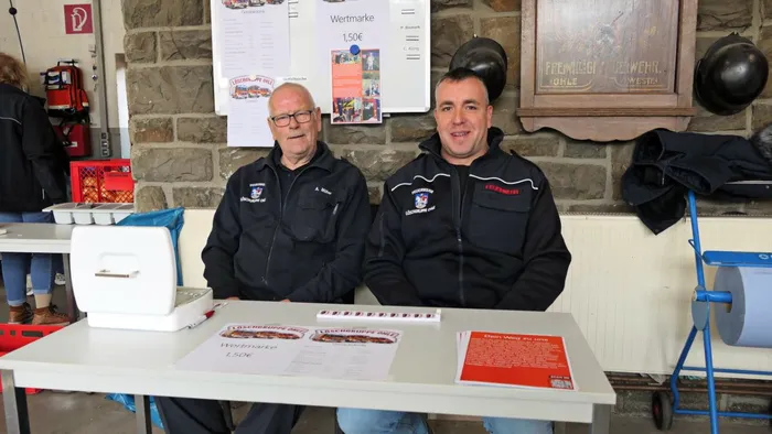 Zwei Männer in schwarzer Feuerwehruniform sitzen an einem Tisch mit Werbematerialien vor einer Steinmauer. Im Hintergrund hängen Plakate und ein Schild mit der Aufschrift „Freiwillige Feuerwehr Ohle“.