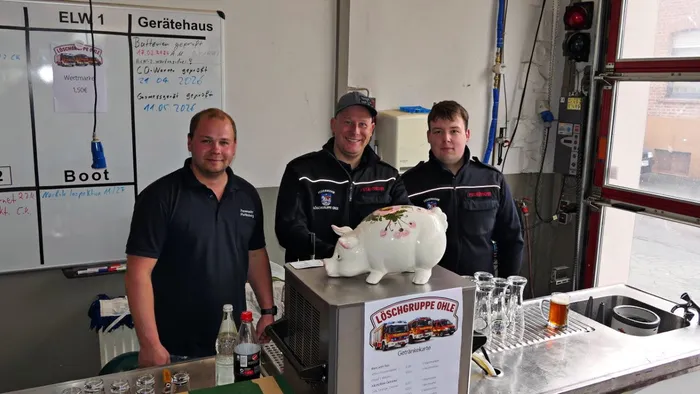 Drei Feuerwehrleute stehen hinter einem Tresen mit Getränken und einer Spardose in Form eines Schweins im alten Feuerwehrgerätehaus.