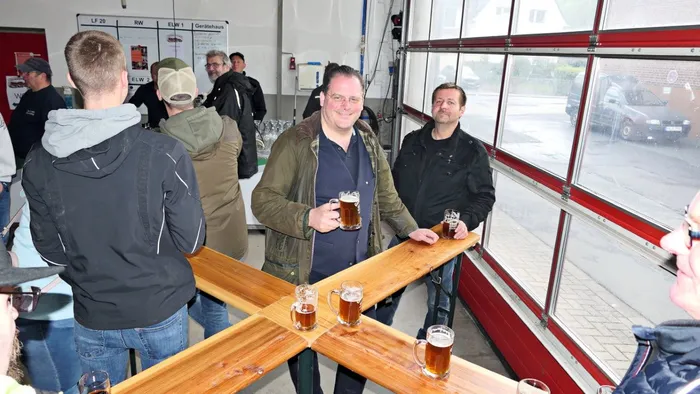 Mehrere Männer stehen an einer Theke mit Biergläsern in einem Raum mit großen Fenstern und Rolltoren. Im Hintergrund sind weitere Personen und ein rotes Feuerwehrauto zu sehen.