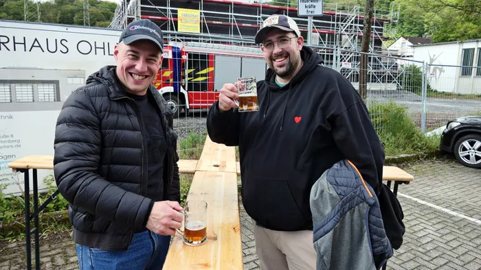 Zwei Männer lächeln in die Kamera und halten jeweils ein Glas Bier in der Hand vor dem alten Feuerwehrgerätehaus Ohle. Im Hintergrund ein rotes Feuerwehrauto und Baugerüste.